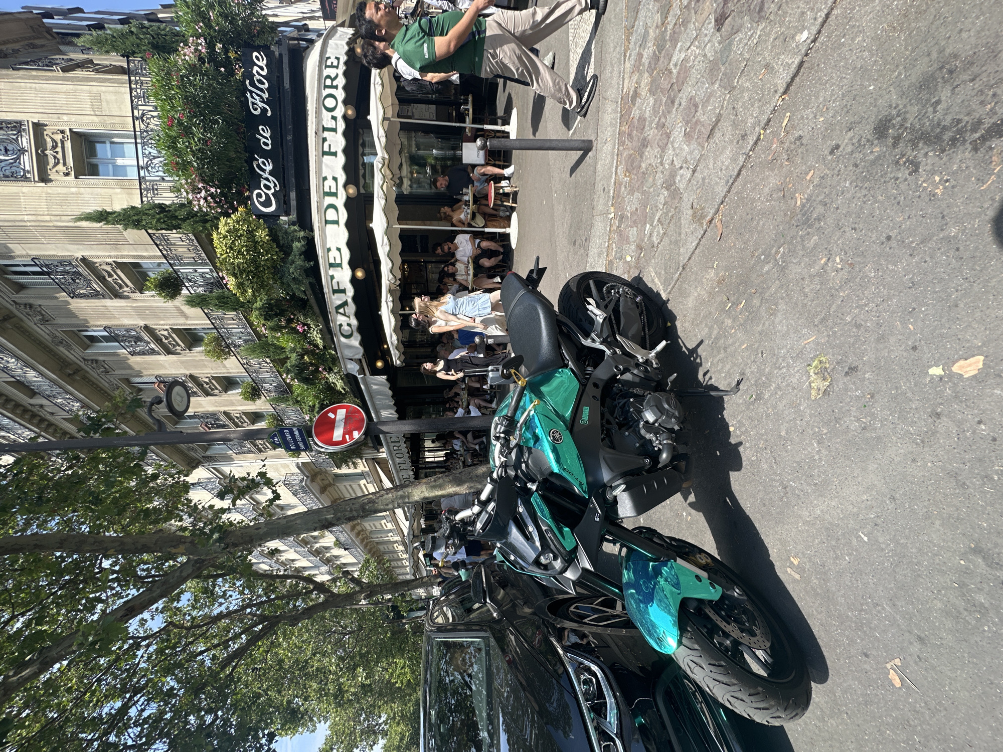 The image depicts a sunny day on a Parisian street, likely in the Saint-Germain-des-Prés district, evidenced by the sign for "Cafe de Flore." The foreground features a vibrant, teal-colored motorcycle parked on the sidewalk. To its left, a portion of a black car is visible, with its headlights and front wheel showing. In the background, the iconic "Cafe de Flore" building is prominent, with its distinctive signage and awnings. Several patrons are seated at tables outside the cafe, enjoying the weather. People are walking along the sidewalk, some carrying luggage. Trees line the street, providing shade, and Parisian architecture with balconies and intricate facades serves as the backdrop. A street lamp and a "no entry" sign are also present. The scene captures a typical urban Parisian atmosphere with a blend of modern elements like the motorcycle and classic cafe culture.