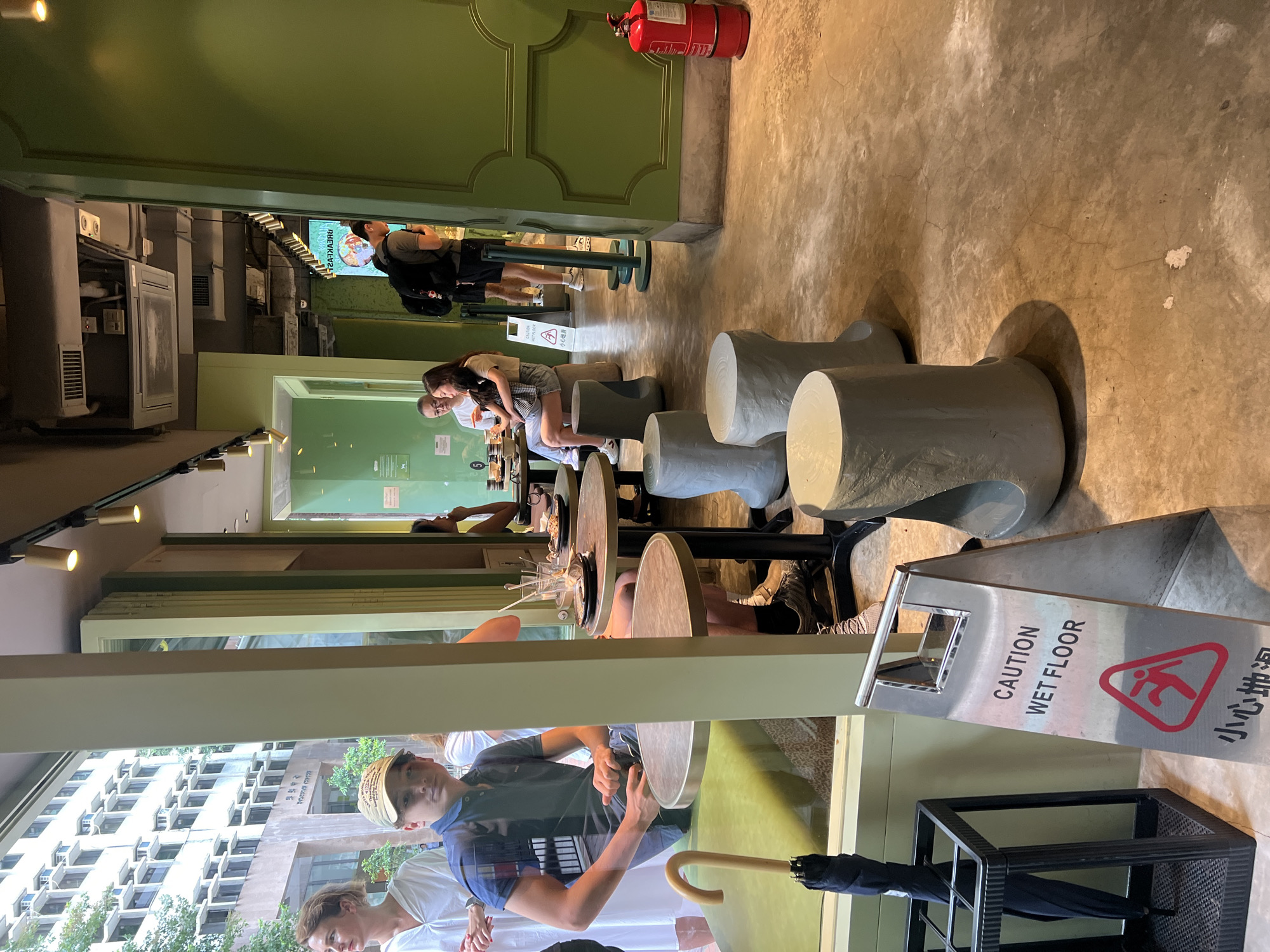 This is an indoor scene likely in Hong Kong, China, depicting the interior of a cafe with a view of an exterior building. In the foreground, to the left, a young man wearing a blue polo shirt and a baseball cap is seated at a round table. An umbrella is stored in a black stand to his left, with a "CAUTION WET FLOOR" sign positioned in front of it. The cafe has green walls and a concrete floor. Several round, grey stools are placed around tables. In the background, a red fire extinguisher stands near a green wall. People are visible dining and conversing at tables, with a woman holding a child, and another man with a backpack walking away. The exterior view shows a modern building with numerous windows and air conditioning units.
