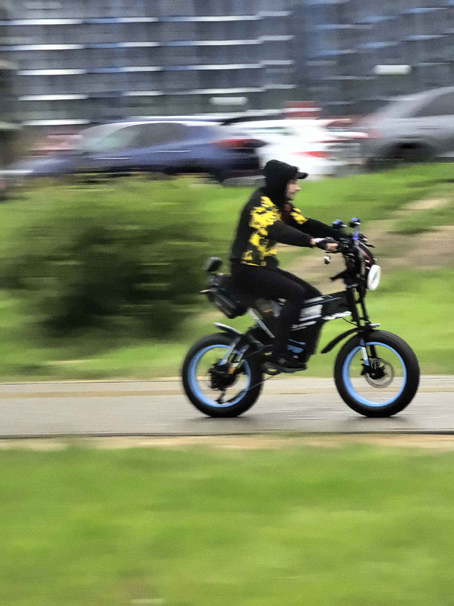 An individual wearing a black hooded sweatshirt with yellow accents and black pants rides a black electric bicycle with blue wheel rims on a paved surface. The bicycle has a white circular headlight and a black seat. The rider is looking to the right, and their hands are on the handlebars. In the background, blurred cars and a building are visible. The foreground consists of a blurred green grassy area. The image appears to be taken with a motion blur effect, suggesting movement.