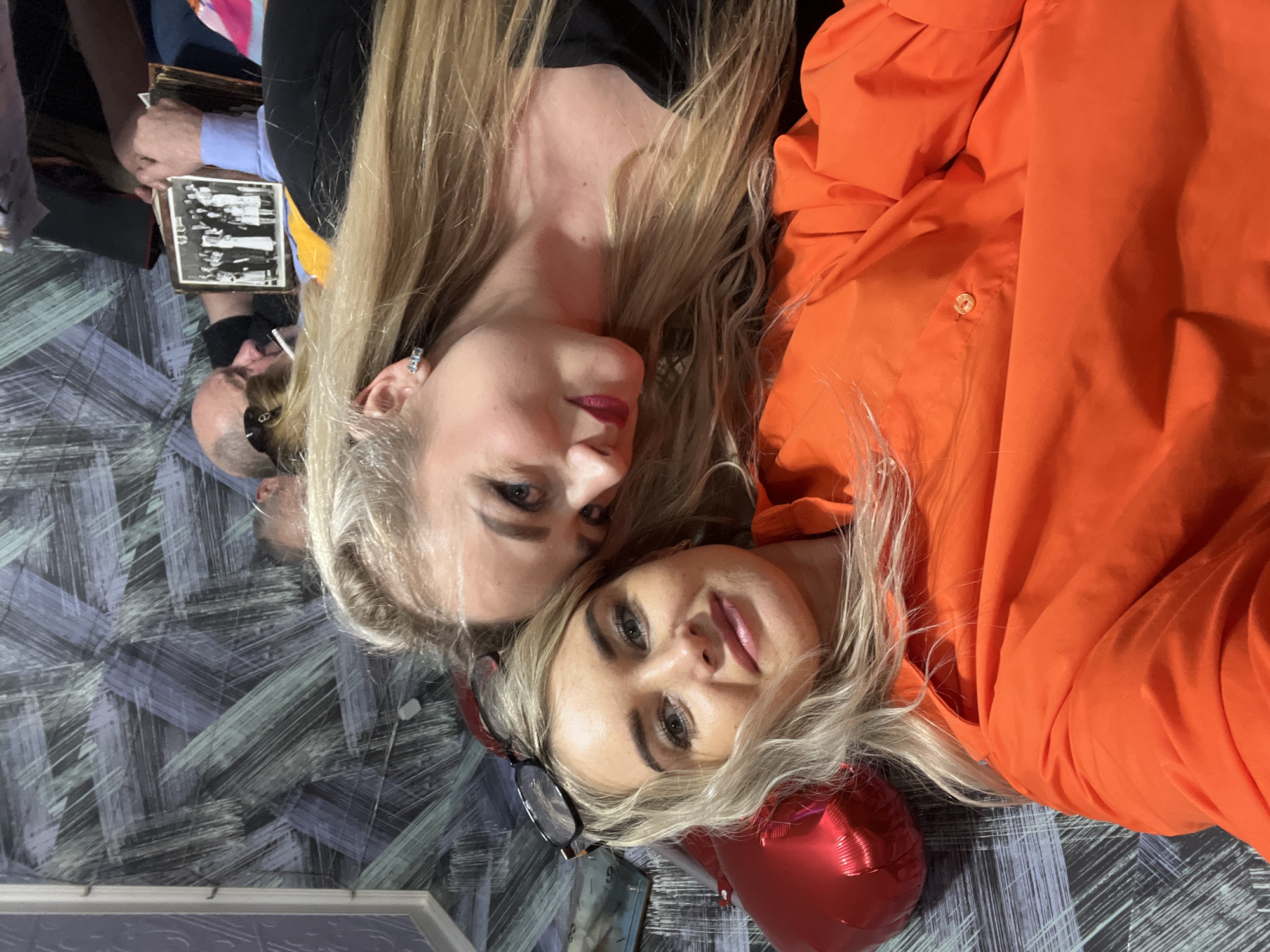 Two blonde women are lying on a patterned floor. One is wearing a black top, the other is wearing a bright orange shirt. Glasses rest next to the woman in orange, and a red heart-shaped balloon is nearby. In the background, other people are visible, including a person holding a framed black and white photo. The floor has a painted pattern of blue and grey. The location is assumed to be Chaikovsky, Russia.