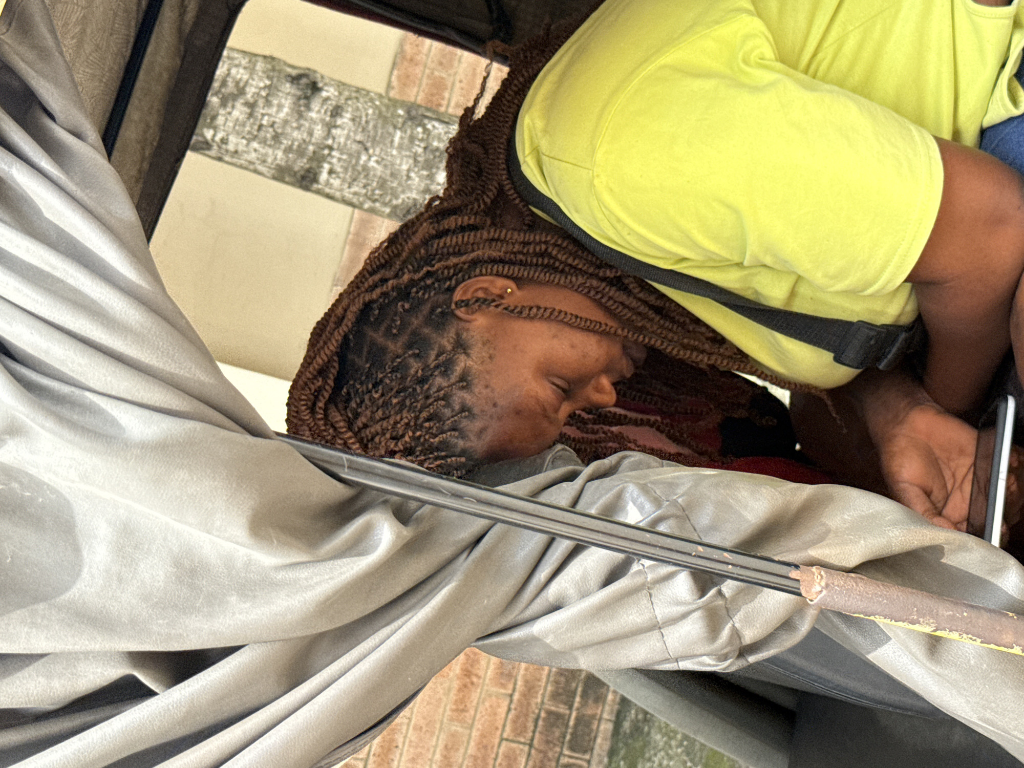 A person is inside a vehicle with a light gray vinyl or leather-like interior. The person has brown braided hair. They are wearing a bright yellow shirt with a black strap across their chest. Their head rests against the interior wall. They have an earring in their left ear. Their hand holds a cell phone. Outside of the vehicle, a brick exterior wall is visible, indicating an urban setting. The location is Ado, Nigeria.