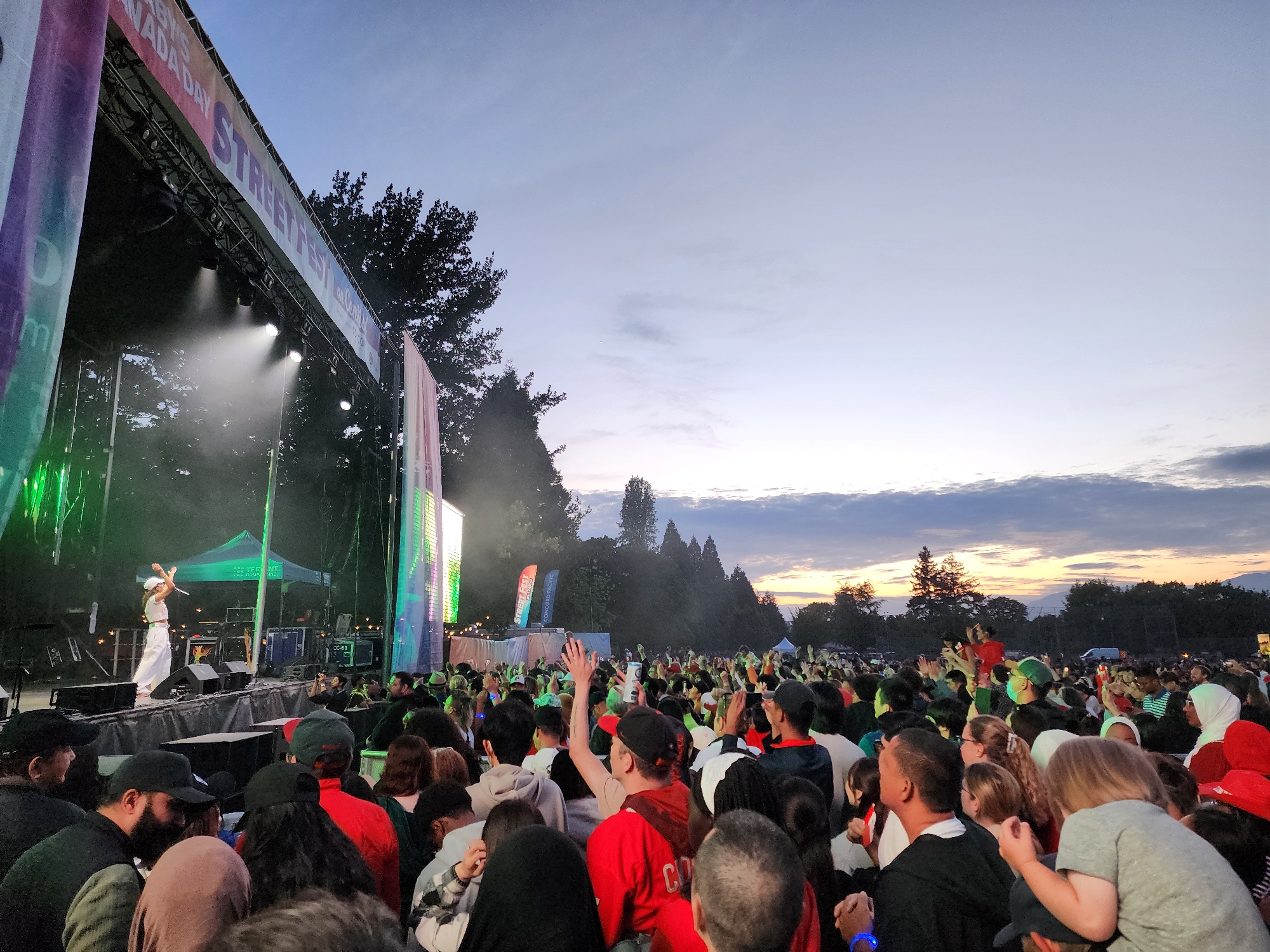An outdoor music festival is in progress at dusk, with a large crowd gathered in front of a stage. The stage has bright green and pink lighting. A performer in white is on stage, gesturing with their hands raised. In the background, trees and a dusky sky with clouds are visible. The crowd is diverse, with many people holding up their hands, suggesting they are enjoying the performance. Some individuals are holding drinks. The overall atmosphere is energetic and festive.