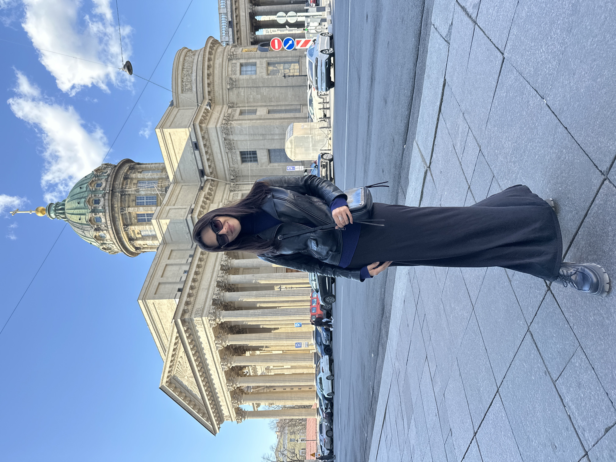 A full-length photograph captures a woman standing on a grey paved sidewalk on a sunny day, with the iconic Kazan Cathedral in Saint Petersburg, Russia, forming the backdrop. The woman, positioned on the right side of the frame, is dressed in a black leather jacket over a dark blue turtleneck, a long dark skirt or dress, and grey and white athletic shoes. She has dark hair and wears dark sunglasses, with a small black shoulder bag slung across her body. She appears to be posing for the camera.
Behind her, the grand neoclassical Kazan Cathedral is a prominent feature, showcasing its distinctive green dome topped with a golden spire and a magnificent semicircular colonnade of tall, light-colored columns. To the woman's left, a street runs parallel to the cathedral, lined with several parked cars, including a silver SUV. Various traffic signs are visible along the street, including red circular signs with a white horizontal bar (indicating 'No Entry' or similar prohibitions) and blue circular signs with a red diagonal line (denoting 'No Stopping' or 'No Standing'). A white 'P' symbol indicating parking is also visible on another sign further down the street. The sky above is a clear blue with a few scattered white clouds, suggesting bright daylight, likely midday or early afternoon. Overhead wires are also visible against the sky.