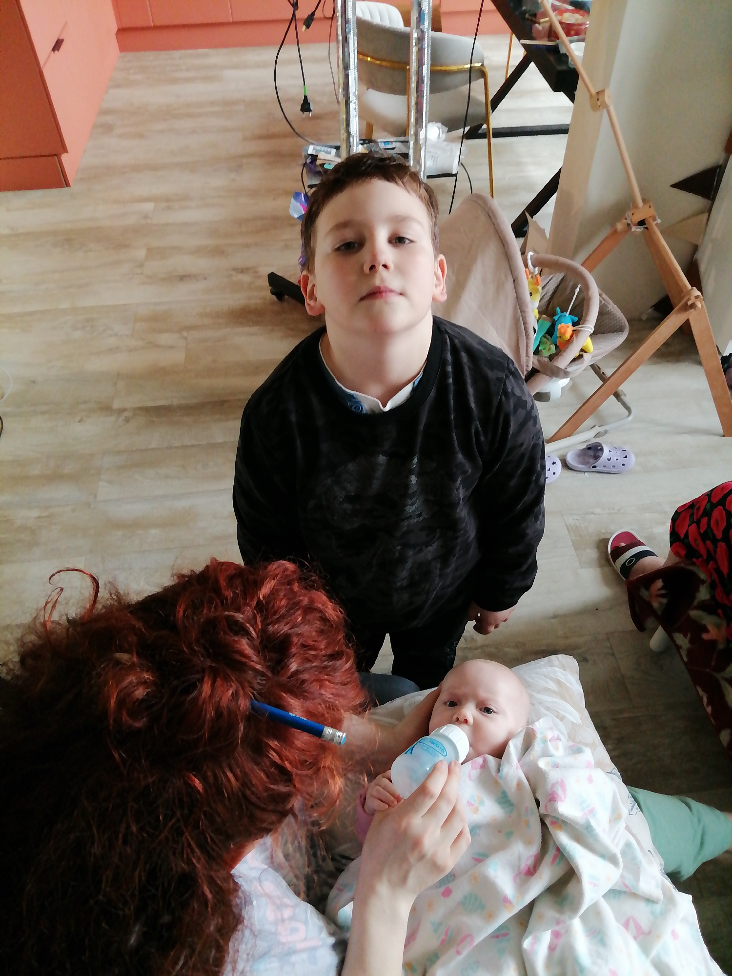 A young boy stands in the foreground of an interior space in Unknown, Unknown, looking directly at the camera with a neutral expression. He is wearing a black patterned sweater. Behind him and to the right is a baby in the arms of an adult woman, presumably being bottle-fed. The woman's face is mostly obscured, but she has red curly hair with a blue pencil in it. The baby is wrapped in a white blanket with a light-colored print and rests on a pillow. In the background, there are chairs, a baby bouncer, a teepee-style structure, and various items, indicating a domestic interior setting. The flooring is light-colored wood-look laminate.