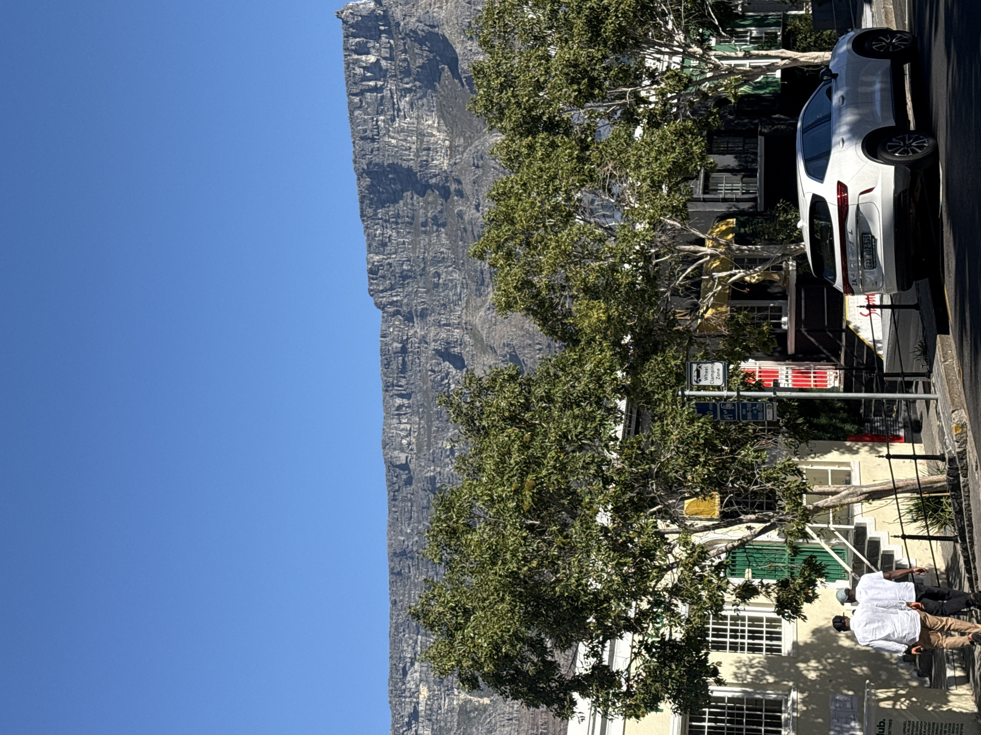 The image captures a street scene in Cape Town, South Africa. Table Mountain dominates the background under a clear blue sky. A white car is parked on the side of the road. Trees line the street, partially obscuring buildings. Two men, both wearing light-colored shirts and darker pants, are walking on the sidewalk. A signpost with multiple signs is visible near the trees. The buildings are multi-storied with visible windows and some have green window frames.