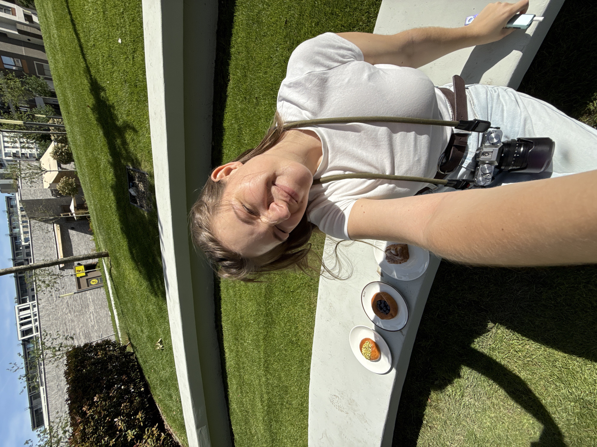 A young woman with light brown hair, wearing a white t-shirt and light blue jeans, lies on a concrete bench in Belgrade, Serbia, taking a selfie. A camera hangs from her neck. Three small white plates with pastries are on the bench beside her. The background includes green grass, trees, modern buildings and a blue sky.