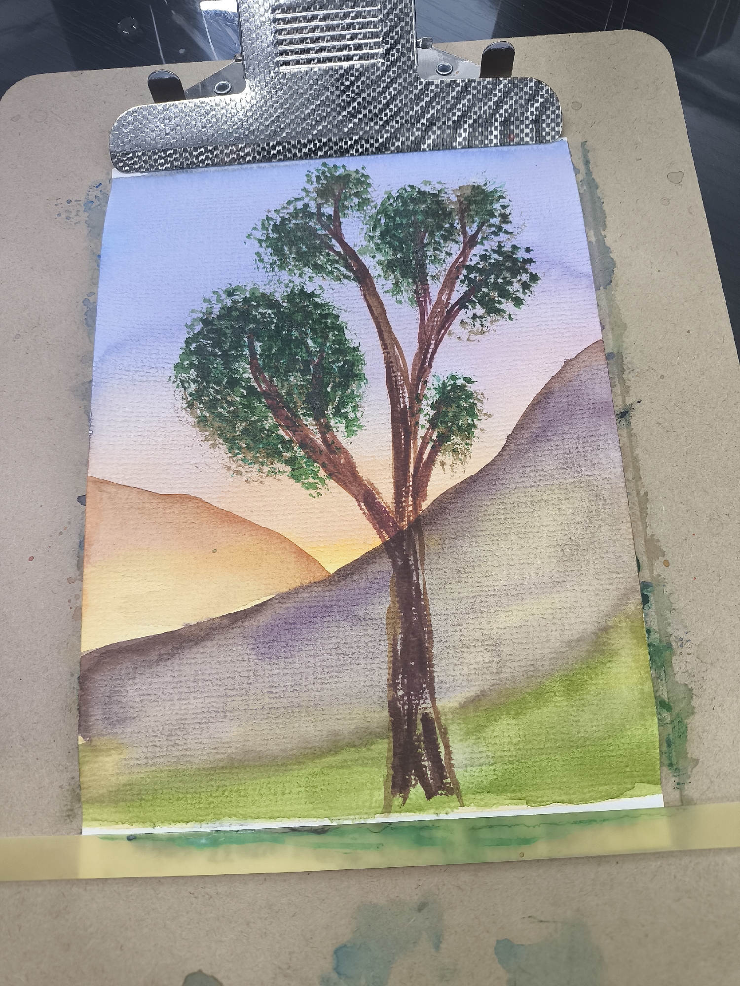 A watercolor painting, depicting a landscape with a tree in the foreground against a backdrop of hills and a sunset. The sky transitions from light blue to soft orange. Two rounded hills are visible on the left, rendered in orange and yellow hues. A larger hill, rendered in shades of gray and purple, occupies the right side. A tree with a brown trunk and green foliage dominates the composition, positioned in front of the hills. The ground is depicted with green watercolor washes, indicating grassy areas. The painting is mounted on a clipboard with a metallic clip at the top. There is a yellow tape lining the bottom part of the painting.
It is not possible to determine the location of this painting based on the provided image. It could be inspired by any location that features trees, hills, and sunsets. There is no clear evidence indicating Moscow, Russia.