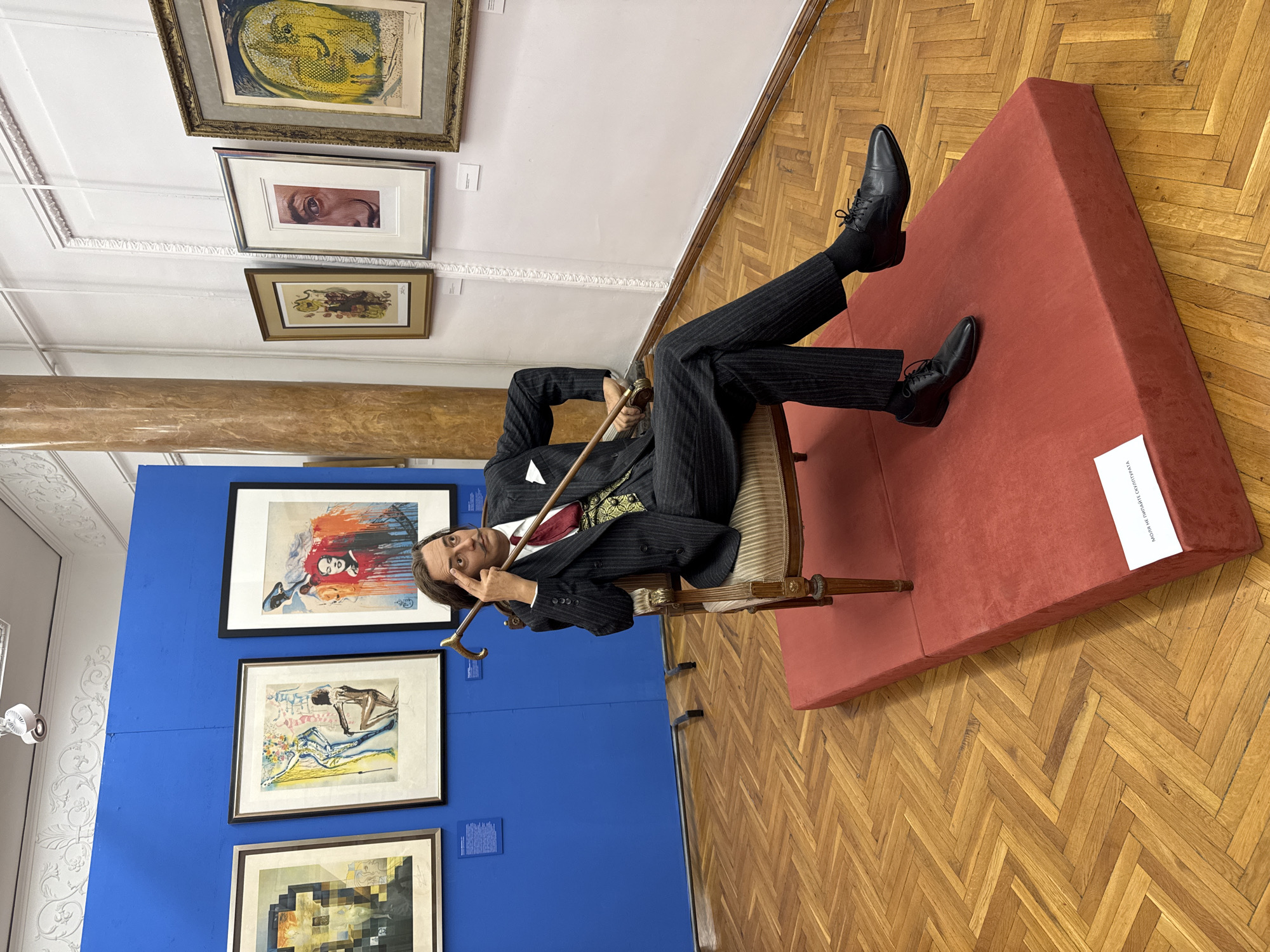 This is a full, eye-level shot of a wax figure of Salvador Dalí sitting on a chair in an art gallery. The wax figure is dressed in a pinstripe suit, red tie, and a decorative vest. He is holding a cane and has his index finger pointed to his temple. The figure is placed on a red platform in front of a blue wall displaying several framed artworks. To the right of the figure, a white wall features more framed art. The floor is made of polished wooden planks arranged in a herringbone pattern. The background includes a large pillar and architectural details on the walls and ceiling. A sign in Cyrillic is visible on the red platform. The location is the Museum of Socialist Art in Sofia, Bulgaria, but the sign indicates it is the National Gallery, Pernik Branch.