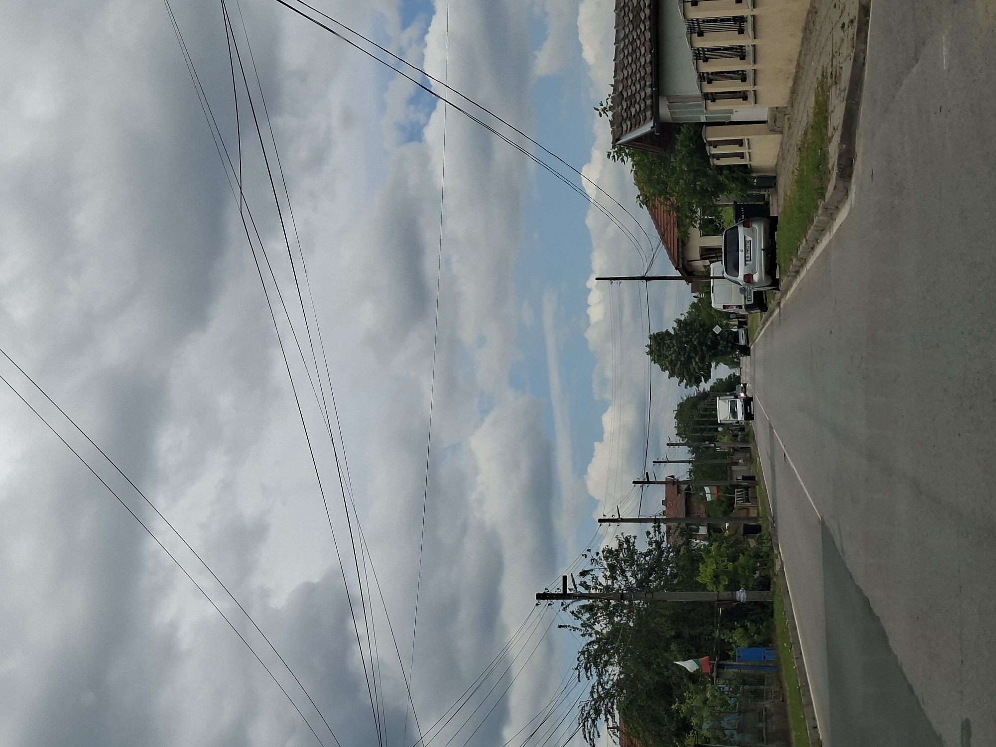 A grey, overcast sky with scattered white clouds looms over a paved road in the town of Detelina, Bulgaria. Overhead, a dense network of power lines crisscrosses the sky, supported by numerous utility poles. The road is flanked by houses with tiled roofs and fenced yards. Several cars are parked along the roadside, including a white van and a silver hatchback. Bulgarian flags are visible on poles outside some of the houses. The scene depicts a typical residential street in a Bulgarian town on a cloudy day.