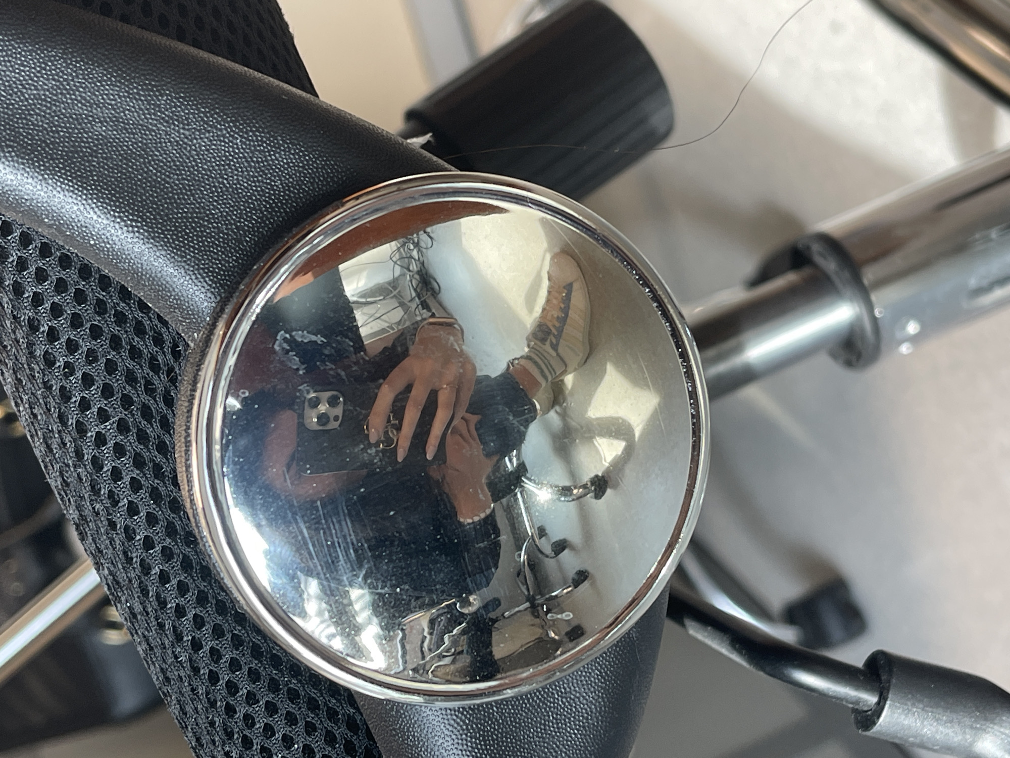 A close-up photograph focuses on a chrome, round swivel joint of a chair, reflecting the photographer, the chair's base, and the photographer's footwear in the polished surface. The chair's upholstery, featuring black faux leather and breathable mesh, dominates the left side of the frame. To the right, a blurred cylindrical metal component of the chair's mechanism is visible. The reflected footwear in the swivel joint appears to be beige and white sneakers. The photographer, captured in the reflection, is holding a phone. The location is unspecified, and no evidence indicates that this image was taken in Sofia, Bulgaria.
