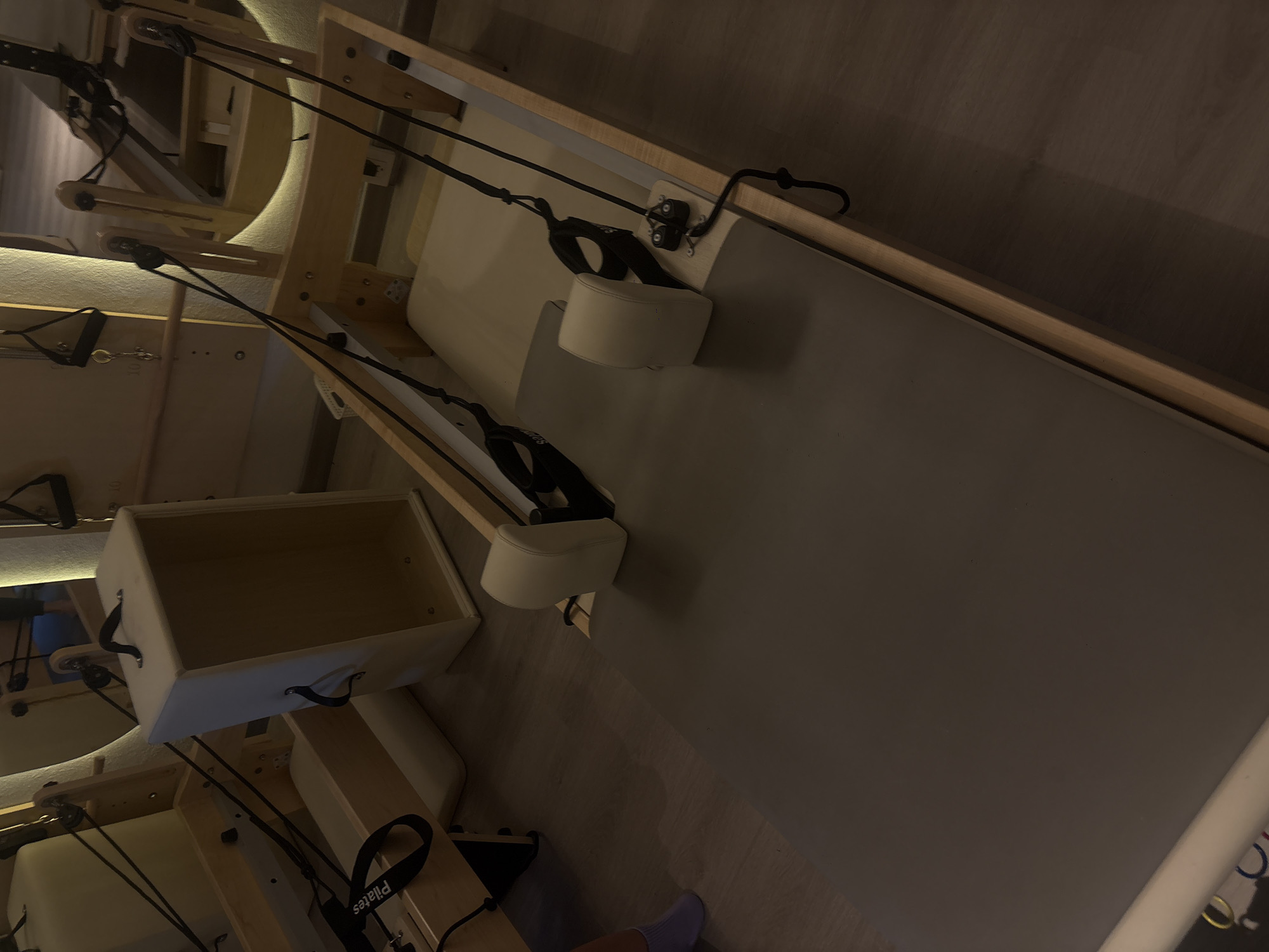 The image depicts a room, likely a Pilates studio, as suggested by the presence of Pilates equipment. The equipment is beige and appears to be made of wood and metal components, with padded surfaces and ropes attached. The room has a wall with a light-colored wood trim. At the lower right, a person's foot is visible. The image appears to be taken indoors, under dim lighting. The exact location within Mexico City cannot be determined from the visual information provided.