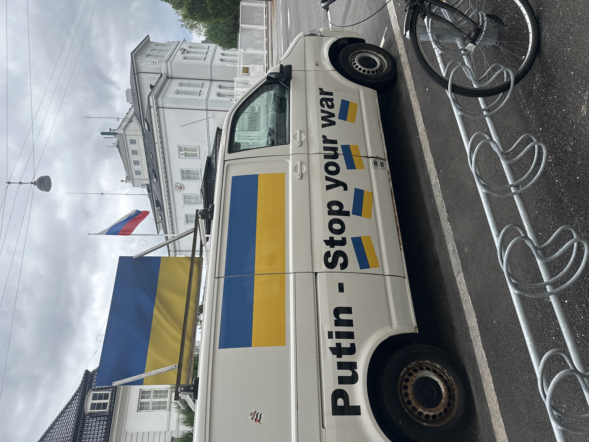 In Copenhagen, Denmark, outside the Russian Embassy, a white van is parked on the street. The van is adorned with the Ukrainian flag on its side and on a panel mounted on its roof. The words "Putin - Stop your war" are prominently displayed in black lettering on the side of the van, accompanied by smaller Ukrainian flags. A Russian flag flies from a flagpole in front of the embassy building. The scene is captured on an overcast day, with a bicycle rack and a bicycle visible in the foreground.