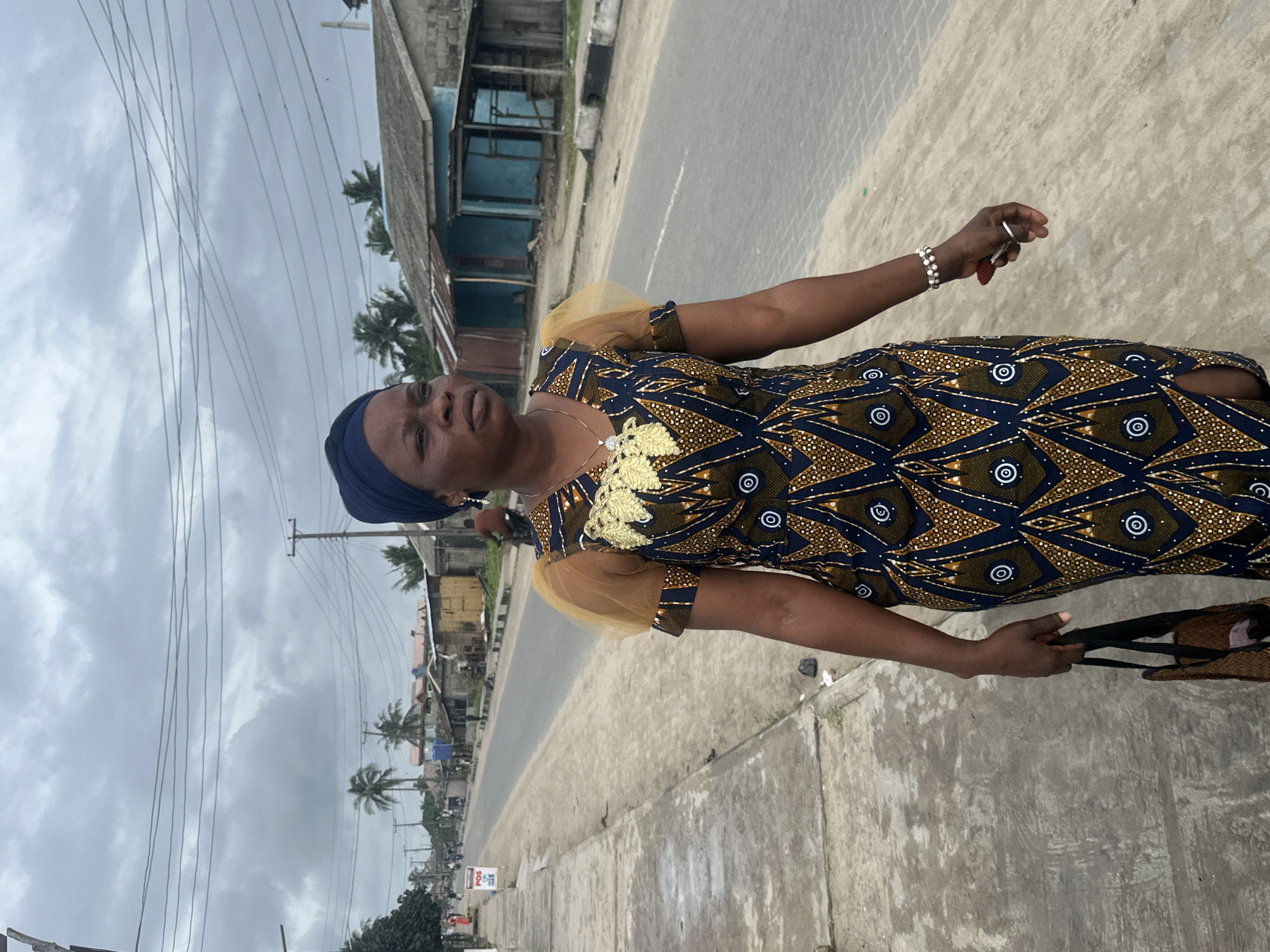 A woman with dark skin and a dark blue headwrap walks along a paved sidewalk beside a road. She wears a patterned dress with a blue, yellow, and white geometric design, and her sleeves are made of sheer yellow fabric. She is carrying a dark woven bag in her left hand and is holding a small red object in her right hand. Her jewelry includes a silver necklace with a pendant and a beaded bracelet on her right wrist, along with a ring.
The road is gray asphalt and appears to be a two-lane street. On the right side of the road, there are small, weathered buildings with corrugated metal roofs and some blue painted structures. On the left side of the road, beyond the sidewalk, is a line of palm trees and more buildings in the distance. The sky is overcast with heavy gray clouds. The overall environment suggests a humid and possibly tropical climate.
This scene is captured in the city of Badagry, Nigeria, known for its historical significance and coastal location. The presence of the road, buildings, and palm trees are consistent with the visual landscape of Badagry.