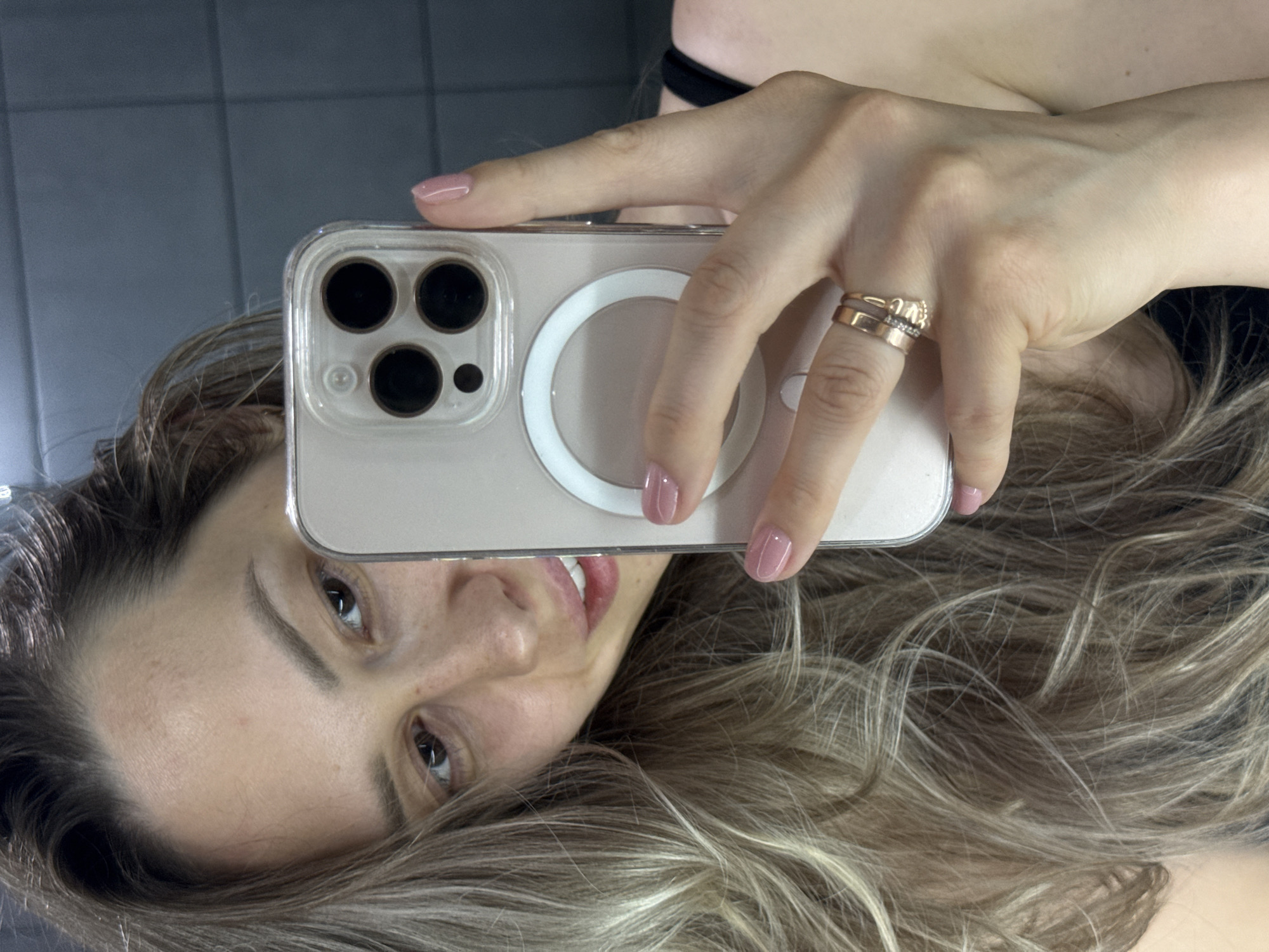 An overhead mirror selfie captures a person with a partially visible face, featuring dark eyes and a slight smile. The individual's light brown hair with blonde highlights cascades around their face and shoulder. They are holding a smartphone in a clear case, showcasing a triple-lens camera system and a white MagSafe ring on its back. The hand holding the phone has light pink painted fingernails. On the person's left ring finger, two gold-toned rings are visible: one is a simple band, and the other is adorned with small, sparkling embellishments. A dark strap, possibly from clothing, is faintly visible. The background consists of a dark, tiled wall with light gray grout lines, suggesting an indoor setting, possibly a bathroom, in Chaikovsky, Russia. The image is rotated 90 degrees clockwise, giving it an unconventional orientation. The activity depicted is the act of taking a casual mirror selfie. No specific text is discernible.