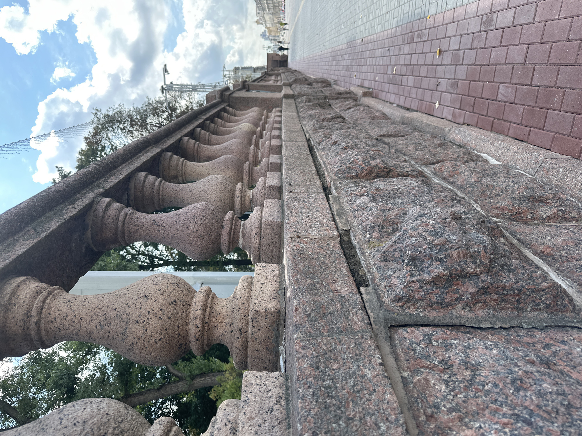 The image depicts a street scene in Minsk, Belarus. In the foreground, a stone balustrade runs along the right side of the frame, characterized by its textured, pinkish-brown granite construction and a series of decorative balusters. The balustrade is set atop a lower wall made of similarly textured granite blocks. To the right of the balustrade, a paved sidewalk is visible, laid with reddish-brown rectangular pavers arranged in a diamond pattern. Beyond the sidewalk, a street stretches into the distance, with a few indistinct figures walking along the pavement and some cars on the roadway. The background shows buildings and trees under a cloudy sky.