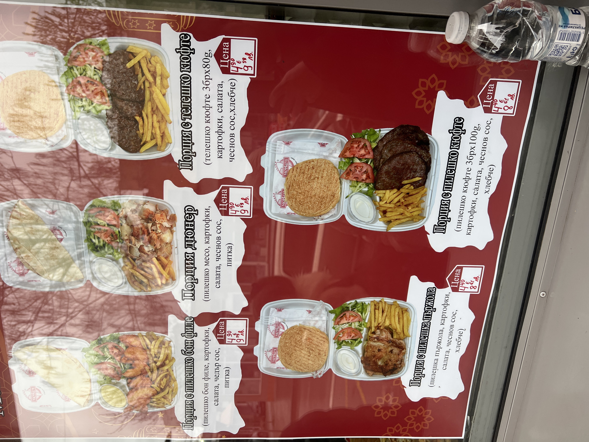 The image shows a menu board for a fast-food restaurant, displaying various meal options. The meals listed, from top to bottom, include "порция с телешко кюфте" (portion of veal meatballs), "порция дюнер" (portion of doner), "порция с пилешко бон филе" (portion of chicken boneless fillet), and "порция с пилешка пържола" (portion of chicken steak). Each meal is described with its ingredients, such as potatoes, salad, and sauces. Prices in both Euro and Bulgarian Lev are listed next to each meal. The photo appears to be taken outdoors, with a water bottle visible in the background. The menu board has a red background decorated with floral patterns. The location is likely in Goce Delchev, Bulgaria, indicated by the use of Bulgarian language and currency.