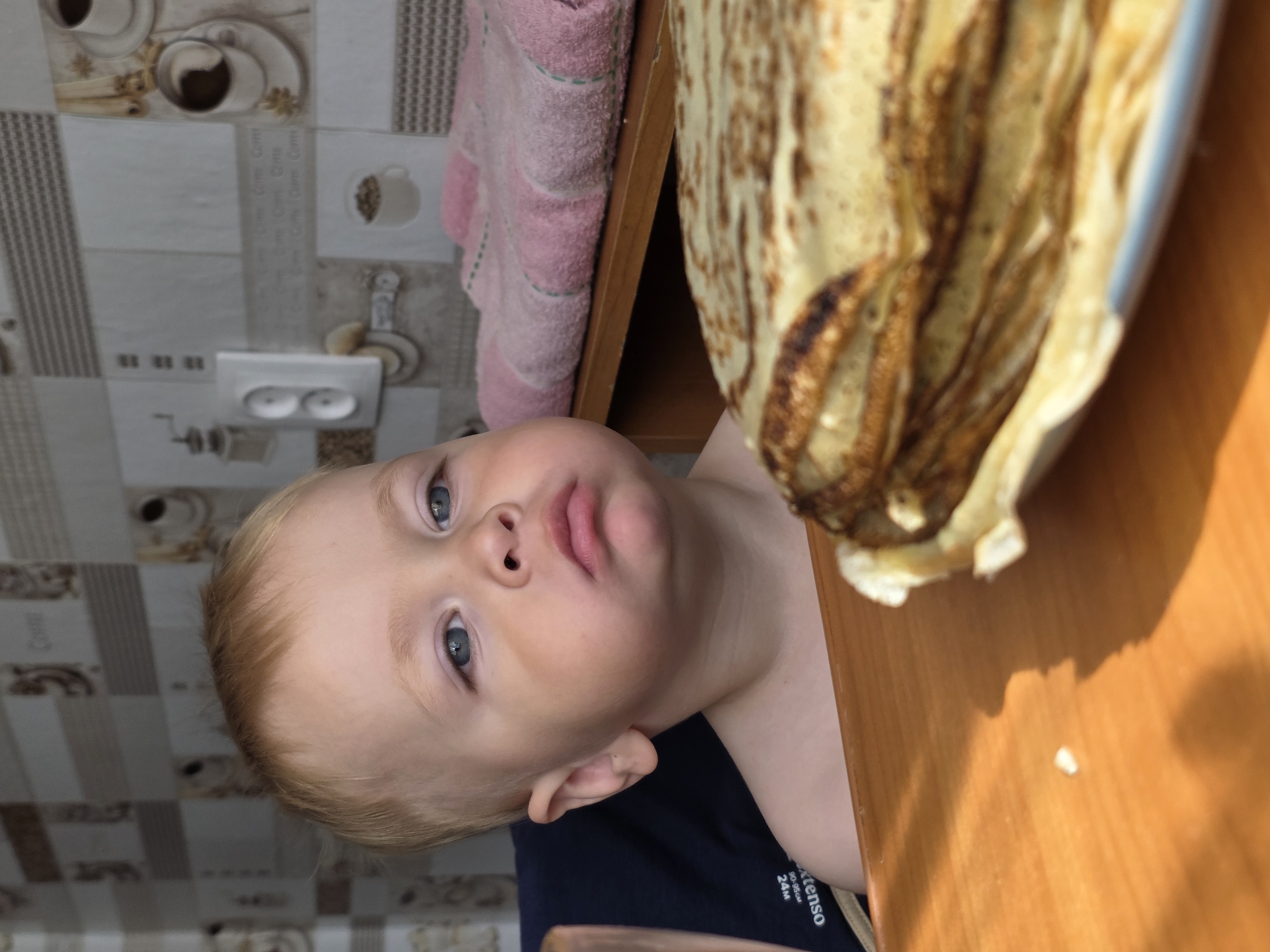 A young child with blond hair and blue eyes is seated at a wooden table, looking to the right of the frame with a slightly pouty expression. In the foreground, a stack of pancakes is partially visible on a white plate. Behind the child, a kitchen wall is decorated with tiles featuring coffee-themed imagery. A pink towel is draped over a shelf or counter to the right. The environment appears to be indoors, likely a home kitchen. The image was taken in the city of Chaikovsky, Russia.