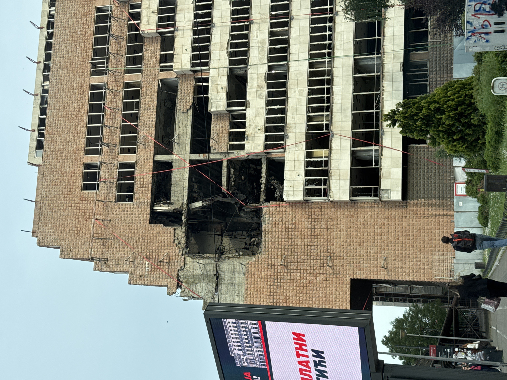 The image shows a damaged building in Belgrade, Serbia. The building has a large section missing, exposing the interior. The exterior is a light brown color with numerous window openings. Some of the window frames are visible. There are red cables or lines hanging from the damaged section.
In the foreground, there is an electronic billboard.
A few people are visible on the sidewalk near the building. A man in a black jacket and blue jeans is standing and looking towards the building. A woman in black is walking nearby.
The environment includes trees and greenery.