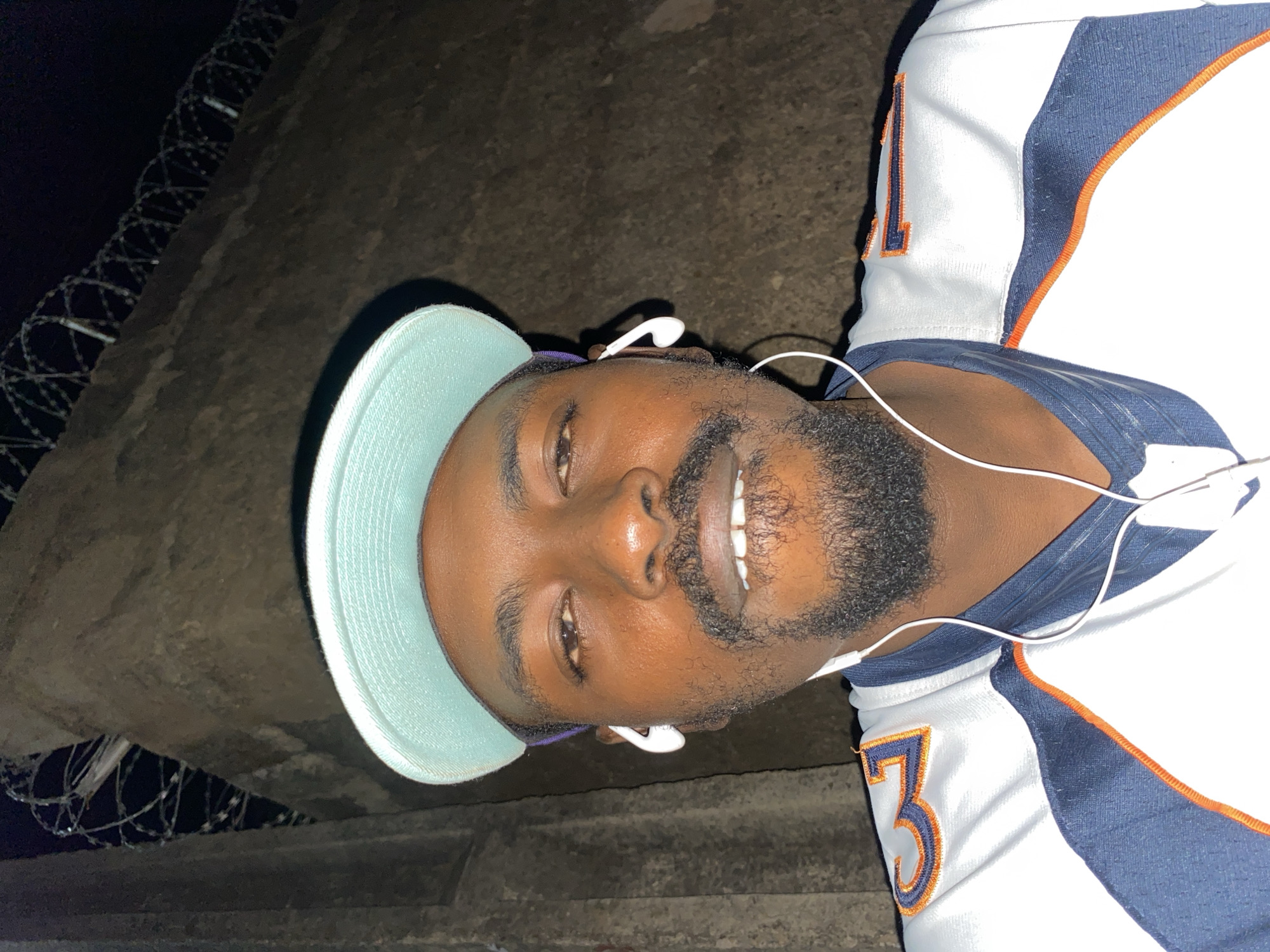 A close-up, rotated portrait shows a man, likely of African descent, looking directly at the camera with a slight smile. He has dark skin, a short beard, and a mustache. He is wearing a light blue baseball cap and white wired earphones, suggesting he is listening to audio. His attire includes a white sports jersey with blue and orange accents, featuring the number '1' on the shoulder and '3' on the side, possibly indicating a team or player number.
The background is dark, indicating the photo was taken at night or in very low light conditions, with artificial illumination on the subject. A prominent feature in the background is coiled razor wire, suggesting a secure boundary, wall, or fencing. The ground or surface beneath the man appears dark and textured, possibly concrete or stone. The overall impression is a casual portrait taken outdoors in an urban or semi-urban setting, possibly in Barakin Akawo, Nigeria, where security fencing like razor wire can be common. The man appears calm and friendly.