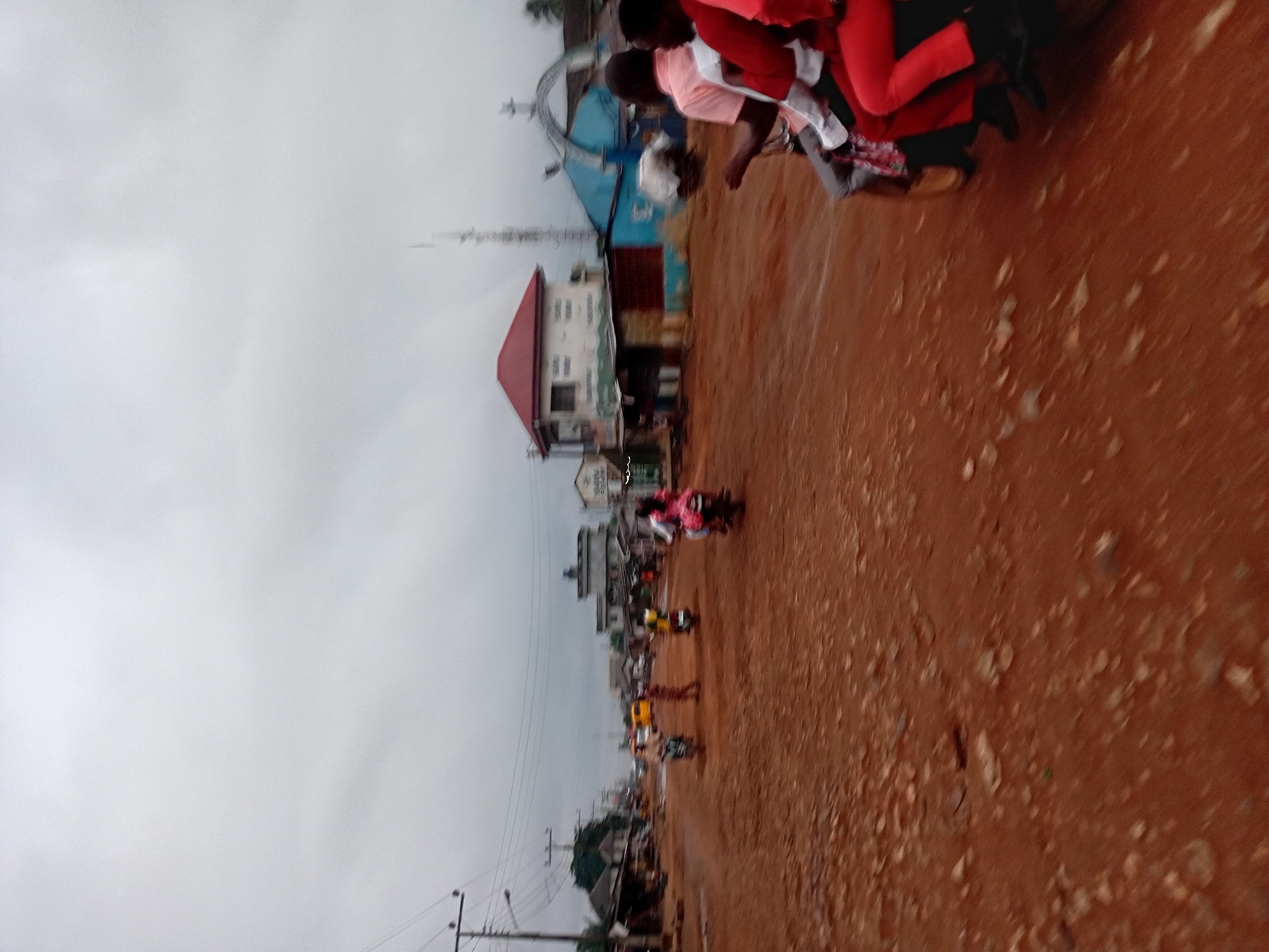 A medium shot captures a bustling scene on a dirt road in a rural or semi-urban setting under an overcast sky. The road surface is a reddish-brown dirt, littered with small stones and debris. Several people are seen on motorcycles, some of them passengers. In the foreground, two people are riding on a motorcycle, the passenger in the rear wearing a bright pink outfit. To the left, other motorcycles and individuals are visible, some appearing to be moving through the scene. Buildings line the sides of the road, featuring various architectural styles and states of repair. These structures include shops, houses, and what appears to be a church with a prominent arched entrance and a cross. A tall telecommunications tower is visible in the distance, and power lines stretch across the upper portion of the frame. The overall impression is one of daily life and activity in a community.