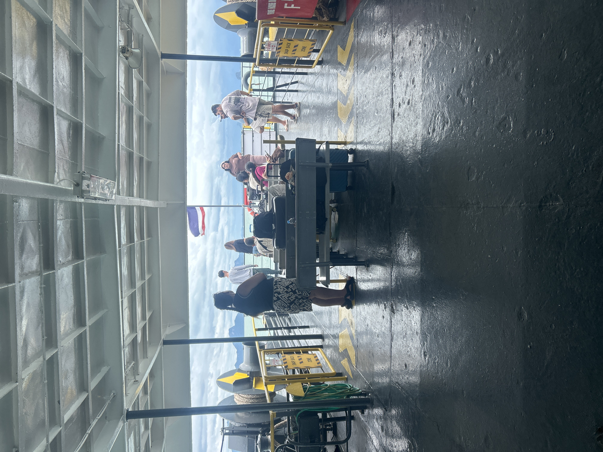 The image depicts the deck of a ferry with several people and luggage, viewed from the rear of the vessel. The deck is wet and reflective, with yellow arrows painted on the surface. In the background, a cloudy sky with mountainous islands is visible. A Thai flag is flying on the left side of the frame. The ferry appears to be moving or docked in a coastal area, likely near Thong Nian, Thailand, given the tropical mountainous landscape. People are standing and sitting, some looking out at the sea, others interacting. Luggage is placed on the deck and on benches. The overall scene suggests a mode of transportation and travel within a scenic, aquatic environment.
