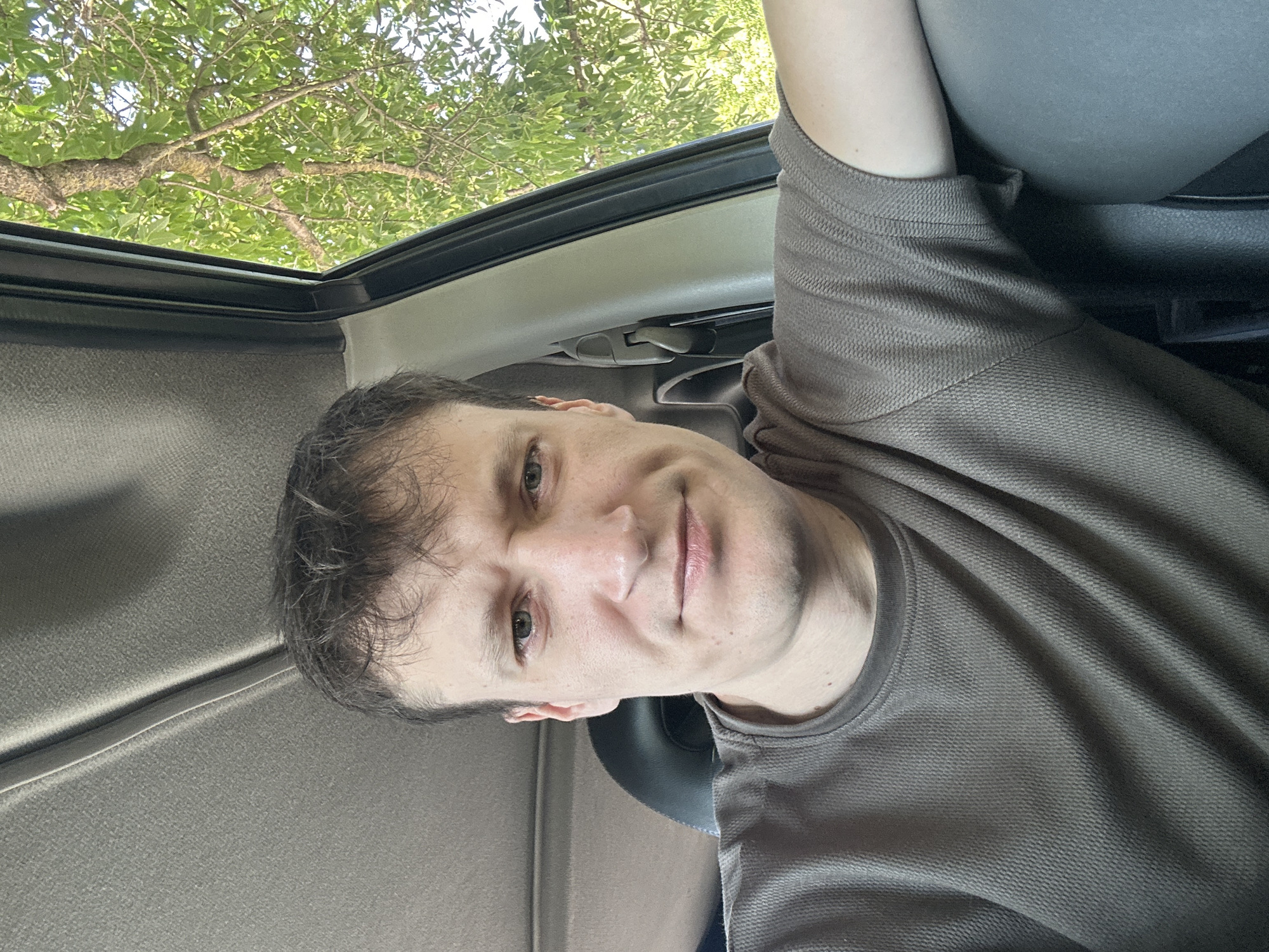 In Makiv, Ukraine, a man with short brown hair and light green eyes is seated in the passenger seat of a vehicle. He is wearing a brown, textured t-shirt. His left arm is resting on the car's interior panel. Outside the open car window, green foliage and tree branches are visible against a bright sky, suggesting a daytime scene, possibly in a rural or park-like setting within Makiv. The interior of the car appears to be a light gray fabric headliner and dark gray seat.
