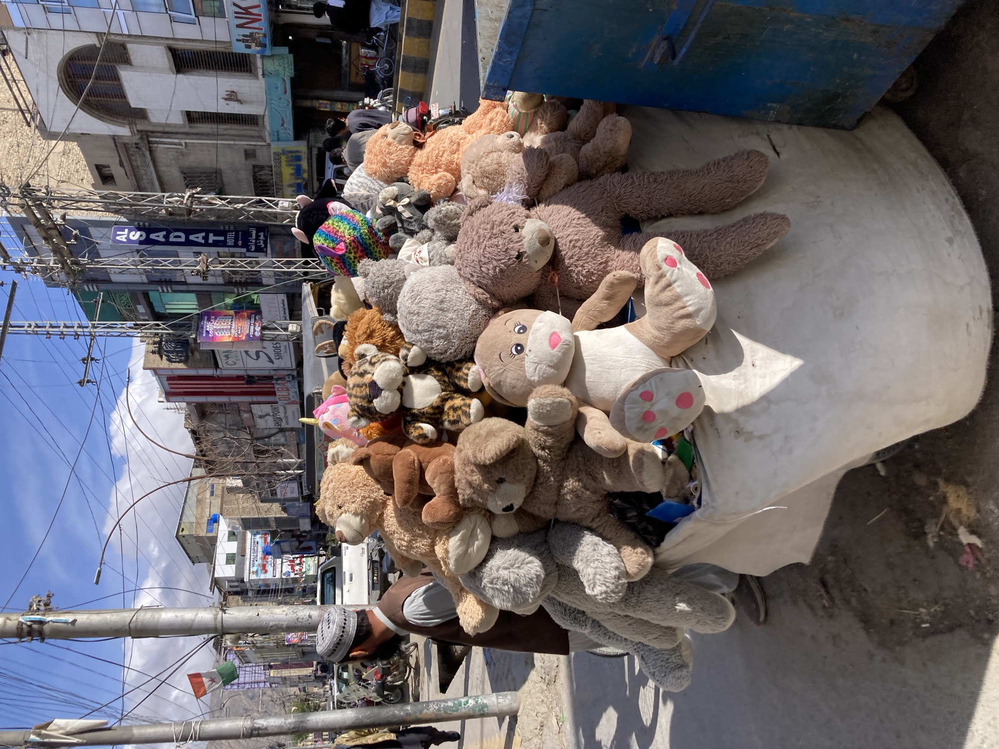 A large pile of stuffed teddy bears fills the cart in Quetta City Tehsil, Pakistan. Various brown, gray, and tan teddy bears are stacked on top of each other, with a white stuffed animal with pink polka dots on its foot. A man wearing a brown vest and a white cap stands next to the cart. In the background, buildings and power lines are visible under a blue sky.