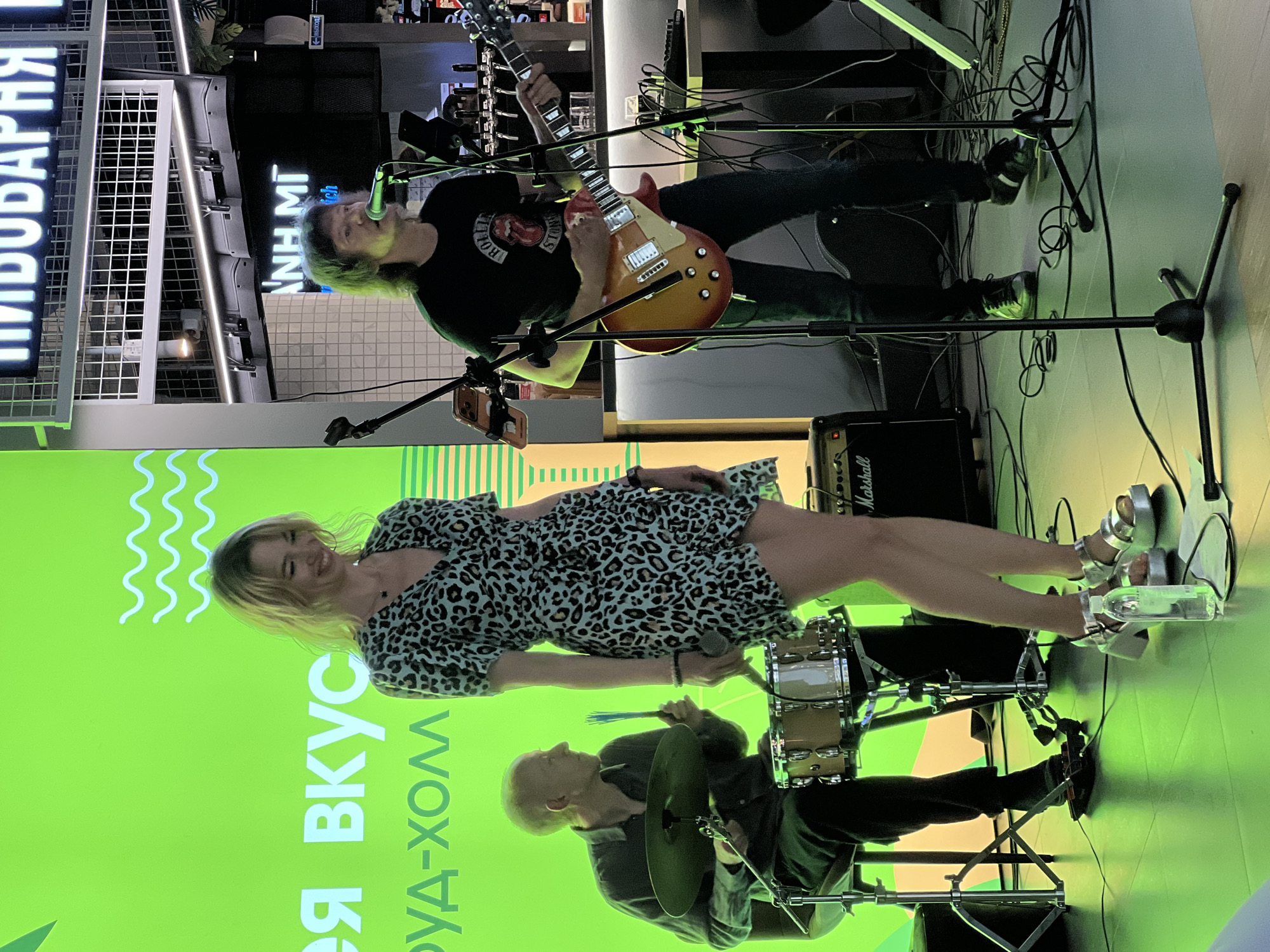 In Ladozhskoe Ozero, Russia, a live musical performance is taking place. A woman with blonde hair, wearing a leopard-print dress and silver platform sandals, stands center stage, smiling and holding a microphone. To her left, a bald man sits behind a drum kit, holding drumsticks. To the woman's right, a man with long, dark hair and wearing a black t-shirt that appears to be a Rolling Stones t-shirt, plays an electric guitar. He is also positioned near a microphone. The background features a bright green screen with white wave-like graphics and Cyrillic text, and overhead, there's a sign with more Cyrillic text. The stage is illuminated with green lighting, reflecting off the tiled floor. Various audio equipment, including amplifiers and microphones, are visible.