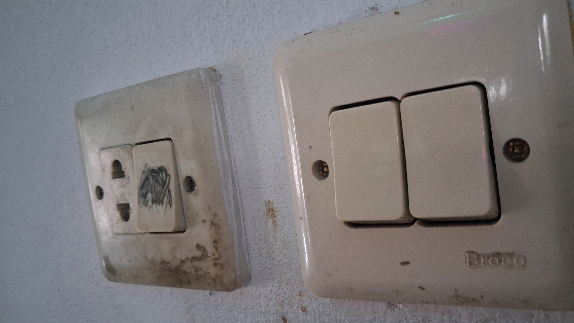 The image shows two electrical outlets on a dirty, textured white wall. The outlet on the left is a universal outlet with three slots, one of which is covered by a switch that has been scribbled on with dark lines. The outlet on the right is a double switch with two rectangular buttons. The word "Broco" is visible on the lower right corner of this outlet. The overall scene suggests an indoor environment, likely a home or building in Giriwarno, Indonesia.