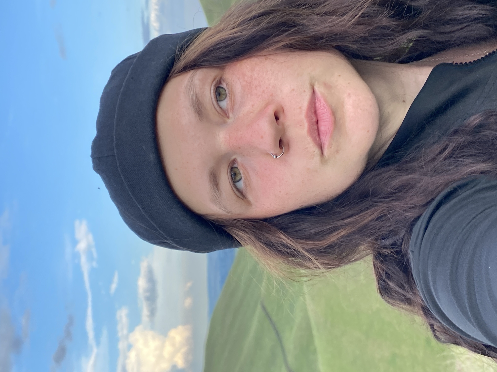 A close-up selfie shows a person wearing a black beanie and a black jacket. The person has brown hair, green eyes, freckles, and a nose ring. In the background, rolling green hills are visible under a blue sky with white clouds. The location is identified as Verkhneozernoe, Russia.
