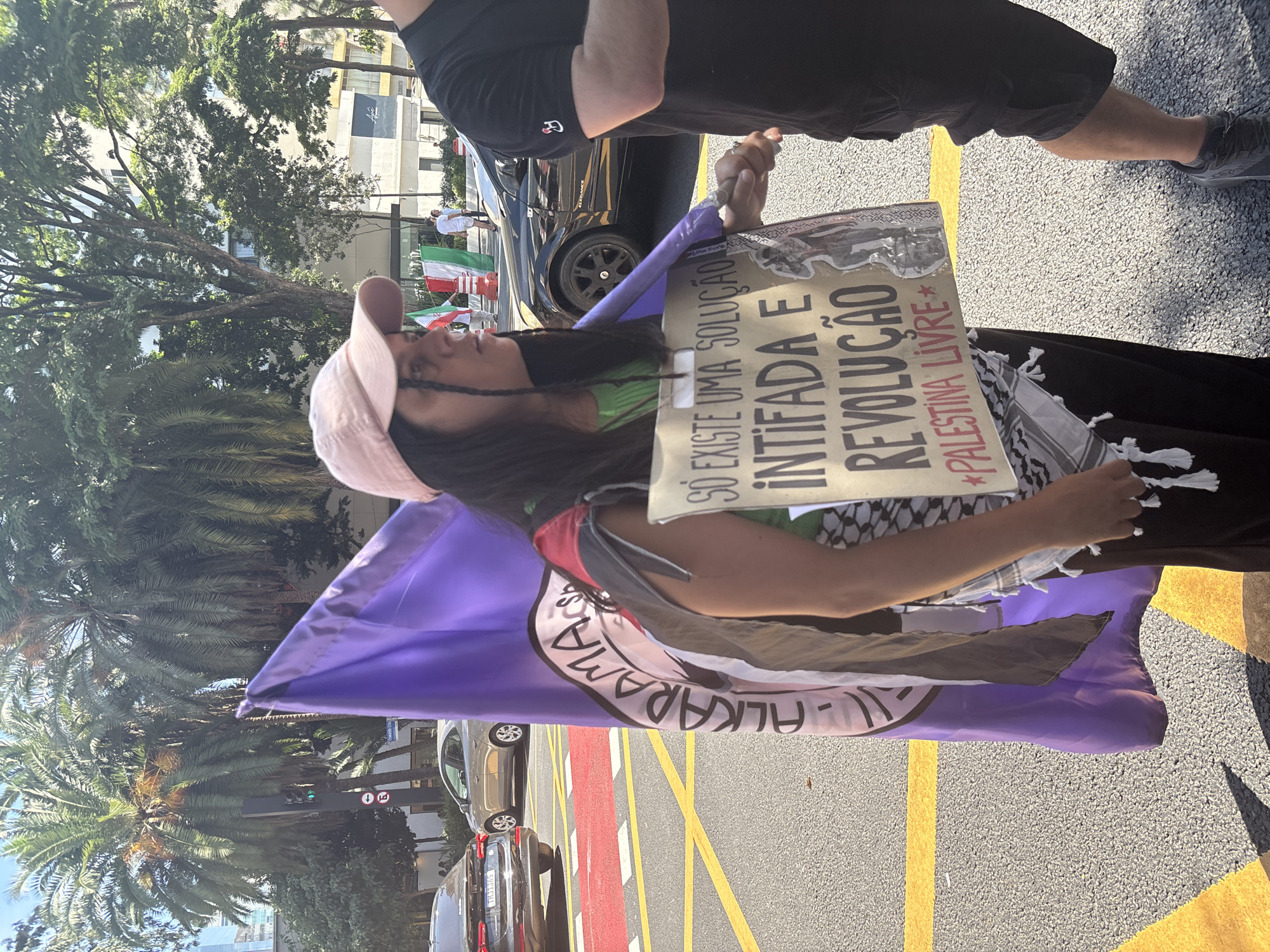 The image shows a woman at a rally in São Paulo, Brazil. She is wearing a pink cap and holding a purple flag and a sign that reads "SO EXISTE UMA SOLUÇÃO INTIFADA E REVOLUÇÃO *PALESTINA LIVRE*" in Portuguese. The flag has the Arabic word "Al-Karama" written on it. The woman is also wearing a keffiyeh scarf. In the background, there are trees, buildings, and other people. Iranian flags are visible in the background. The street has red and yellow paint on it.