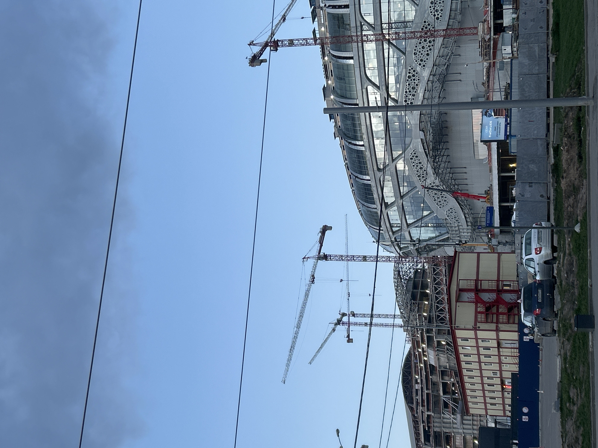 An image rotated ninety degrees depicts a large, modern stadium under construction in Moscow, Russia. The stadium features a distinctive curved, glass-like facade with an intricate, geometric exterior framework, including sections with circular perforations. Multiple tall tower cranes are actively operating around the building, signaling ongoing construction.
Adjacent to the main stadium structure, there is a multi-story building with red and beige paneling, possibly a supporting facility or an annex. In the foreground, a blue construction barrier separates the building site from a road where several cars, including a white hatchback and a dark sedan, are parked. A grassy verge is also visible by the road. Overhead power lines traverse the upper part of the frame.
The sky is a clear, light blue, with some darker clouds visible on the left side. The presence of artificial lights within the partially completed building suggests the time of day is likely late afternoon or early evening.
A blue billboard is visible near the construction site, displaying text in Russian: "ВЫБИРАЙ БУДУЩЕЕ" (Choose the future) and the logo "СБЕР" (Sberbank), along with smaller, less distinct text possibly indicating "НОВЫЕ ПЛАТФОРМЫ ДЛЯ МЕРОПРИЯТИЙ" (New platforms for events).