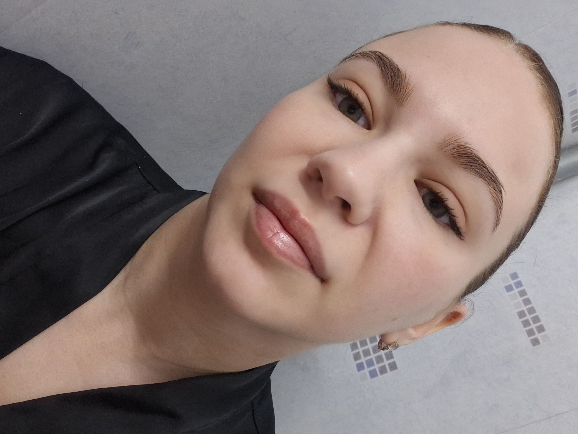 A close-up, angled selfie of a fair-skinned woman with brown hair pulled back, showing the upper portion of her chest and face. She is wearing a black garment. She has carefully shaped eyebrows, dark mascara, and glossy lips. A small earring is visible in her right ear. The background is a light gray wall with a decorative pattern of small blue and white squares. There is no indication of the location being in Chaikovsky, Russia, or anywhere else specific.