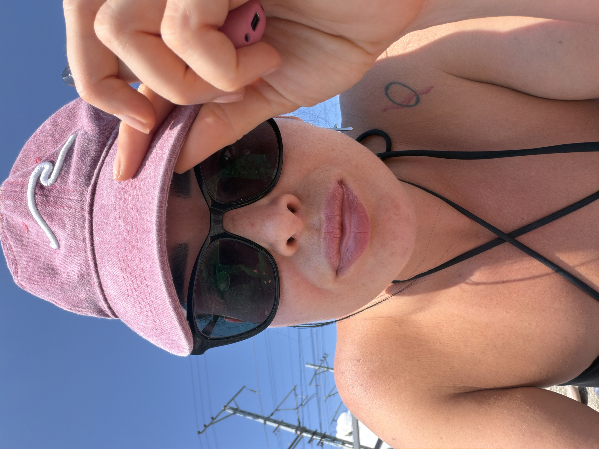 A woman is taking a selfie outdoors under a clear blue sky. She is wearing a pink baseball cap and black sunglasses. Her hand is holding the brim of the cap, and she is also holding a small pink electronic device. She is wearing a black bikini top. The background shows electrical lines and poles. There is a tattoo of a circular design with a line through it on her left shoulder. The scene is bright and sunny, suggesting a warm climate. The image was taken in Sochi, Russia.