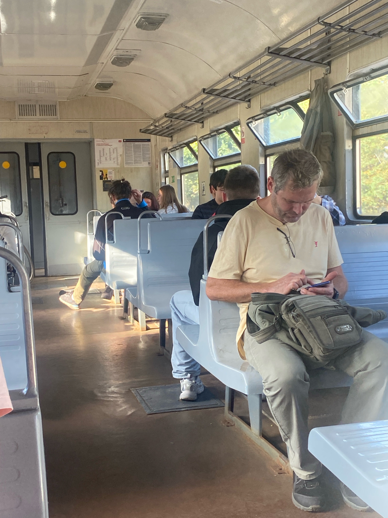 An interior shot of a commuter train in Saint Petersburg, Russia, depicts passengers seated on light-colored plastic benches. The train car has a high, arched ceiling with overhead luggage racks and fluorescent lighting fixtures. The windows are open, allowing natural light to illuminate the scene and offering a glimpse of trees and foliage outside.
In the foreground, a middle-aged man with graying hair is seated, looking down at his smartphone. He is wearing a light yellow t-shirt and khaki pants, and a green utility bag is slung over his shoulder and resting on his lap. His eyeglasses are hanging from the collar of his shirt. To his right, another passenger is partially visible, wearing a blue and white checkered shirt.
Further down the aisle, other passengers are visible, including a young person in a dark jacket and jeans sitting with their legs extended. The floor of the train car is a dark, reflective brown. The overall impression is one of a typical commute on public transportation.
