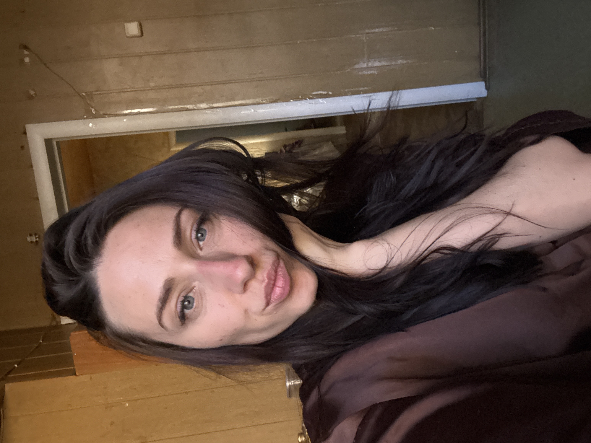 A selfie-style image shows a woman with dark hair and light eyes. She is positioned at an angle, looking at the camera with a slight smile. Her hair is loose and flowing around her face. She appears to be indoors. The background consists of light brown walls and a doorframe, but the location cannot be determined from the provided details. There is nothing in the image to suggest it was taken in Saint Petersburg, Russia.