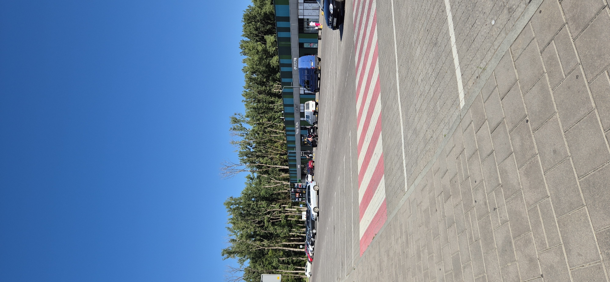 A daytime, eye-level, vertical, wide outdoor shot captures a parking lot and a strip of shops in Drohobych, Ukraine. The sky is a clear, bright blue. In the foreground, a pedestrian crosswalk with red and white stripes is painted on the gray, brick-patterned asphalt. Beyond the crosswalk, a parking lot stretches towards a modern building with a blue and green striped facade. Several cars are parked in the lot, including a silver SUV, a black sedan, and a white van. A blue van is parked in front of one of the shops. A few people are visible in the distance near the entrance of the building, including one person walking with a small child. Trees with lush green foliage line the back of the building, obscuring any view beyond. The scene appears to be a typical commercial area on a sunny day.