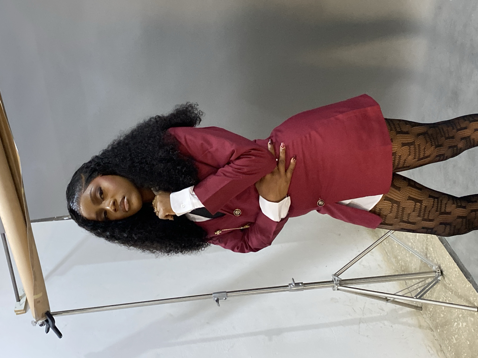 An individual, identified as a woman, stands centered in a studio setting within Gura Top, Nigeria. She has dark, curly hair and wears a maroon blazer-style dress over a white collared shirt with a dark tie. A gold-toned chain with a cross pendant adorns the blazer lapel. Black patterned tights with a geometric design cover her legs. Her right hand is positioned near her chin, while her left hand rests on her waist, and she maintains direct eye contact with the viewer. The background consists of a light grey seamless backdrop, transitioning to a lighter shade on the left, where a metal photography stand and a roll of brown paper are visible. The floor is light-colored concrete.