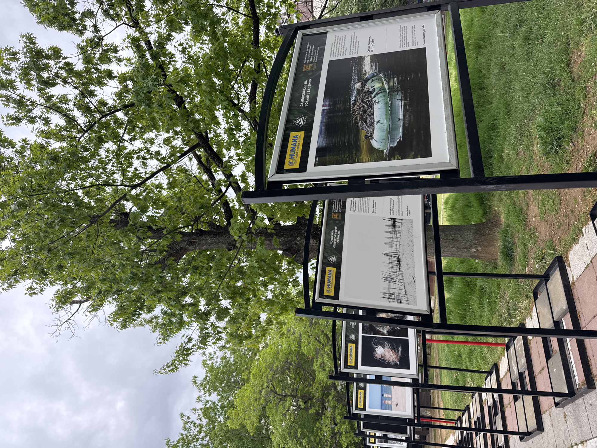 The image shows an outdoor art installation in Blagoevgrad, Bulgaria. Several framed artworks are displayed on a black metal structure. The frames hold images related to environmental pollution and human impact, sponsored by organizations like HUMANA and the municipality of Blagoevgrad. One artwork has the title "ЗАМЪРСЯВАНЕ НА ВОДНИТЕ БАСЕЙНИ" (Pollution of Water Basins). The surroundings include a sidewalk made of red and grey bricks, green grass, and trees, with a large tree with green leaves partially obscuring the sky. The sky is overcast with grey clouds.