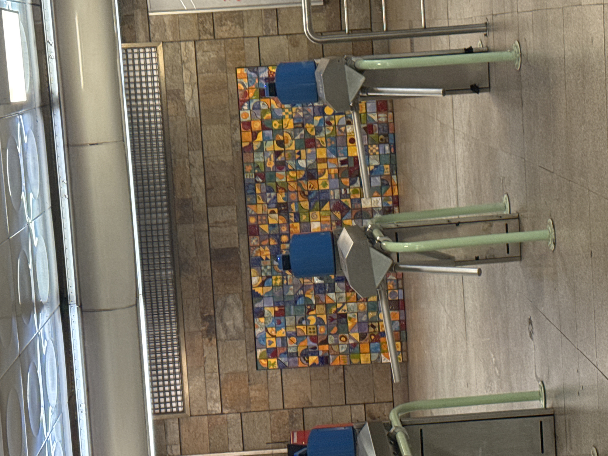 The image depicts an interior view of a subway station. The walls are tiled with rectangular beige tiles and feature an inset panel with a mosaic of small, colorful tiles. The floor is beige and appears to be made of concrete or a similar material. Multiple turnstiles are visible, likely for entering or exiting the station. The turnstiles have blue and gray components and are equipped with green rails. The image appears to be taken in a well-lit area.
The location is likely a subway station in Sofia, Bulgaria, given the presence of turnstiles which are common in metro stations.