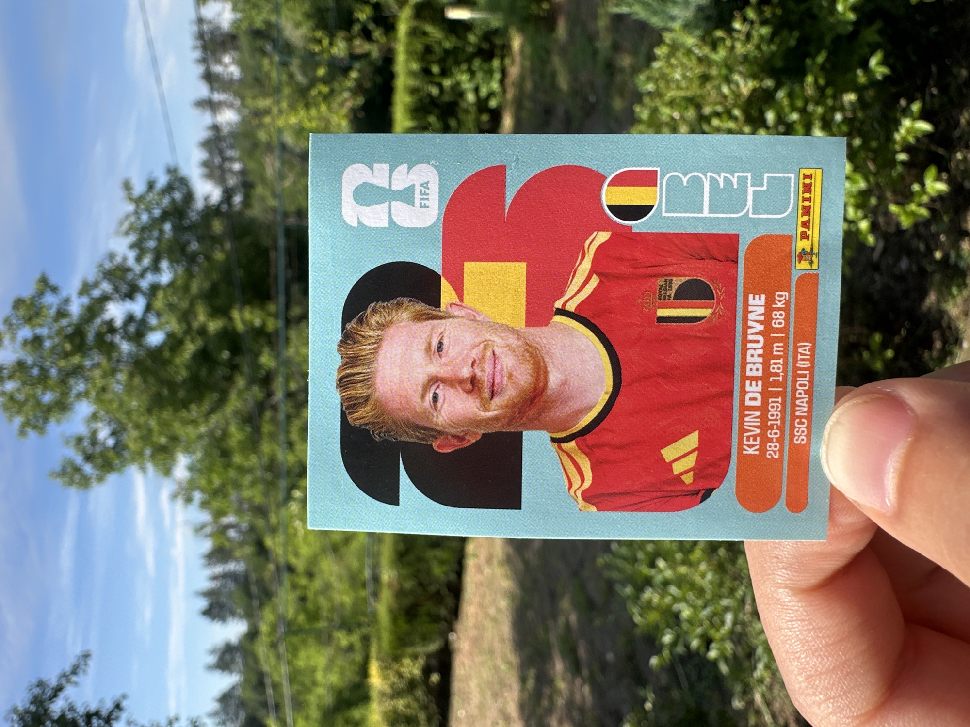 A vertical medium shot shows a hand holding a Panini soccer sticker of Kevin De Bruyne. The sticker is blue with red, yellow, and black graphic elements. De Bruyne, with his signature blonde hair and beard, is depicted in a red soccer jersey with yellow trim. Text on the sticker includes "KEVIN DE BRUYNE," his birthdate "28-6-1991," height "1,81 m," weight "68 kg," and club "SSC NAPOLI (ITA)." The FIFA and Panini logos are also visible. The background is a soft-focus view of green trees and a blue sky with white clouds, suggesting an outdoor setting on a bright, possibly sunny day. The hand holding the sticker is a pale complexion with visible fingernails and fingerprints. The overall impression is that of someone showing off a collectible item outdoors.