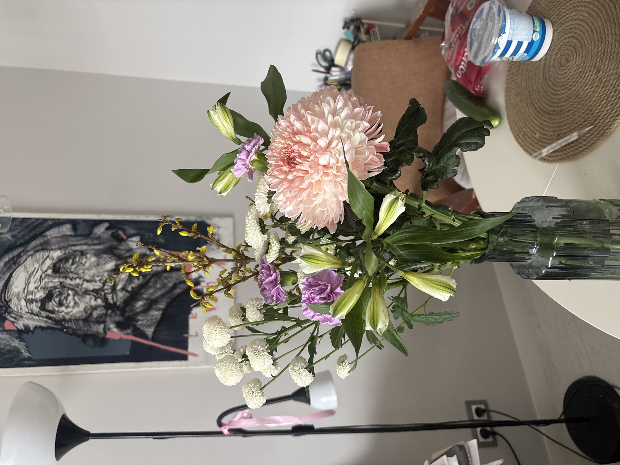 A still life photograph depicts a floral arrangement in a textured glass vase, positioned on a white circular table. The arrangement consists of a large, light pink chrysanthemum as its focal point, surrounded by white pompom chrysanthemums, yellow forsythia sprigs, purple carnations, and unopened white lily buds with green leaves. Behind the table, a beige chair is partially visible, along with a braided placemat, a container of yogurt, a package of food, and a cucumber. In the background, a black and white artwork hangs on a light gray wall, and a modern floor lamp is positioned in the lower left corner. The scene is indoors. The photograph does not provide definitive information about the location being in the city of Krasnoyarsk, Russia.