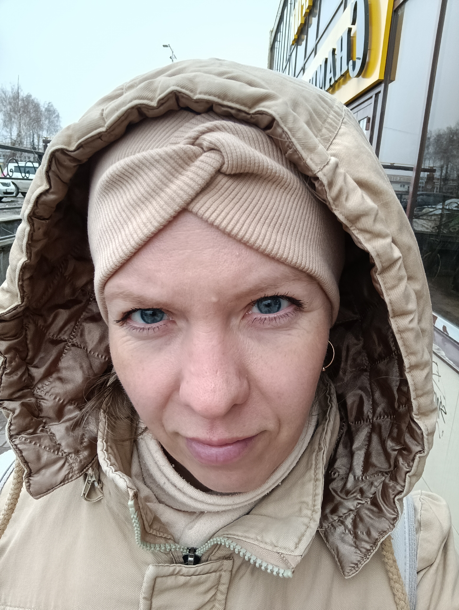 A close-up, eye-level selfie captures a woman outdoors, looking directly at the camera with piercing blue eyes and a neutral expression. She is dressed in warm clothing, including a light beige hooded jacket with a brown quilted lining visible inside the hood, and a matching beige, ribbed, turban-style beanie that covers her forehead and ears. Small gold hoop earrings are visible on her right ear. The jacket's zipper is partially visible.
The scene is an overcast, cool-toned outdoor setting, likely an urban or semi-urban area in Chaikovsky, Russia. In the background to her left, bare trees, a street with parked cars, and a metal fence are visible under a grey sky. To her right, a modern commercial building with large reflective windows features a prominent yellow sign with Cyrillic text. Parts of the sign, specifically "ЧАЙ" and "КОВС", are discernible, strongly hinting at the city name "ЧАЙКОВСКИЙ" (Chaikovsky). The weather appears cloudy and cool, consistent with the woman's layered attire.