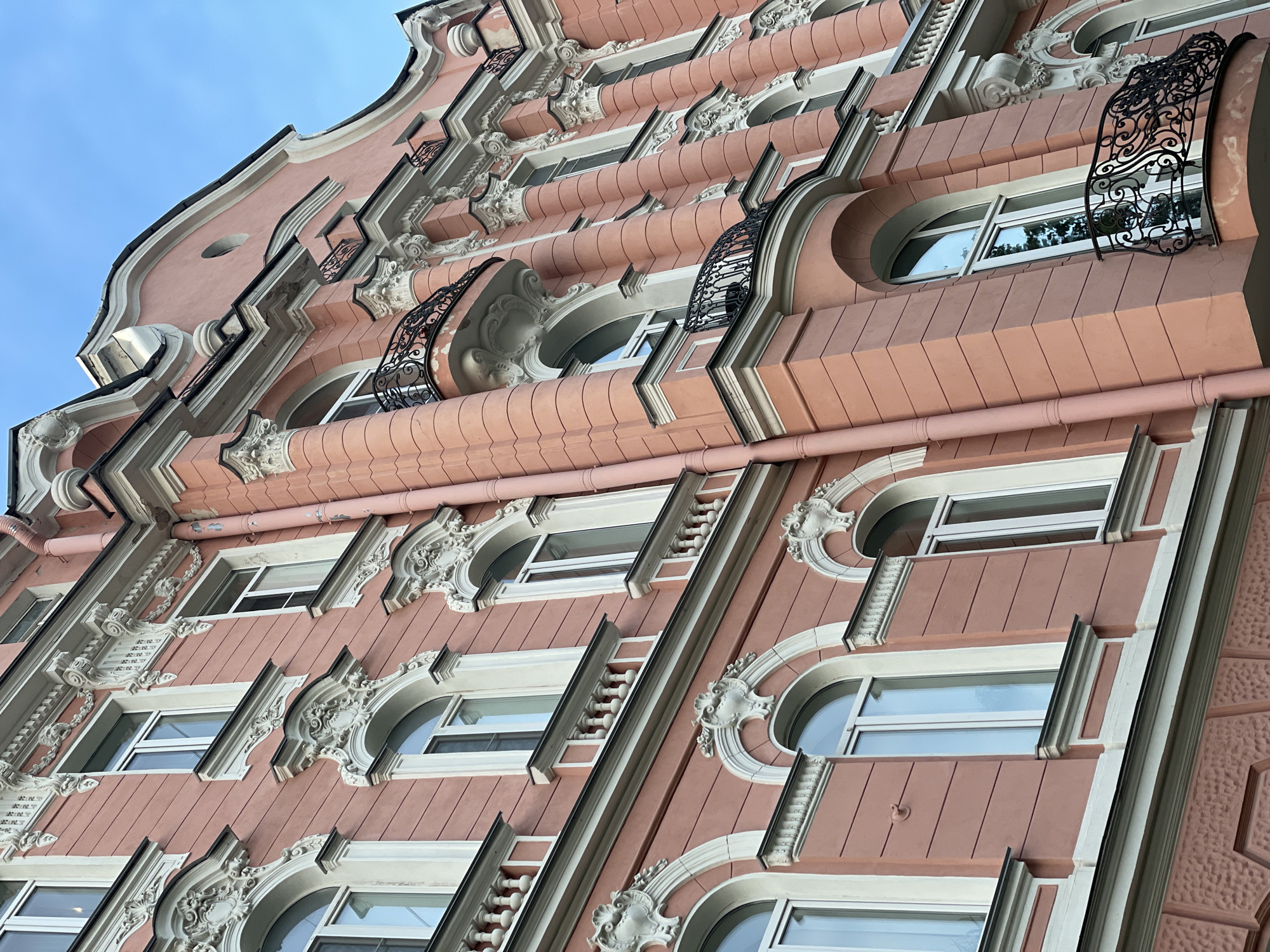A low-angle, upward-looking view captures the ornate, pink-hued facade of a multi-story building against a clear blue sky. The architectural style is Eclectic, featuring elements of Art Nouveau and Neoclassicism. The facade is dominated by numerous windows, primarily arched, with white frames and reflective glass. Many windows are embellished with elaborate, off-white decorative moldings, including volutes, shells, and scrollwork. Some lower windows are fronted by small, white spindle balustrades.
The building's verticality is emphasized by prominent, fluted pilasters, some roughly textured at their bases and smooth above, culminating in ornate capitals. Several small, black wrought-iron balconies with intricate scrollwork project from the facade, including two prominent curved balconies. A large, undulating cornice defines the roofline, against the sky, featuring a small, circular oculus near its peak. A vertical, pink-painted downspout is visible on the right side of the building. The overall color scheme is a dusty rose or pinkish-orange, accented by white architectural details. No individuals are present in the scene.
This building is the House of A.P. Veselov, located at Bolshaya Konyushennaya Street, 10, in Saint Petersburg, Russia.