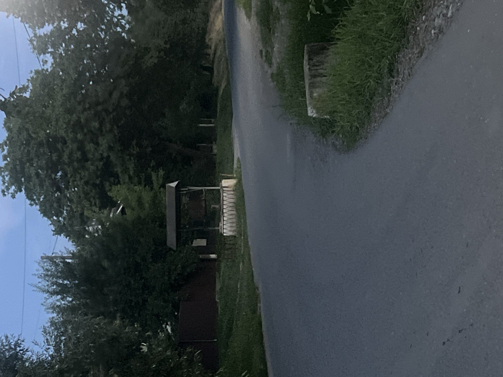 A paved road curves through a sparsely populated area with trees and grass. On the left side of the road, a dark brown fence encloses a property. A covered well, made of stone and wood, stands near the road. A tree stump covered in grass is visible on the right side of the road. The sky is a deep blue, suggesting either early morning or late evening. There are no people visible in the image. The scene appears to be a rural or semi-rural environment.