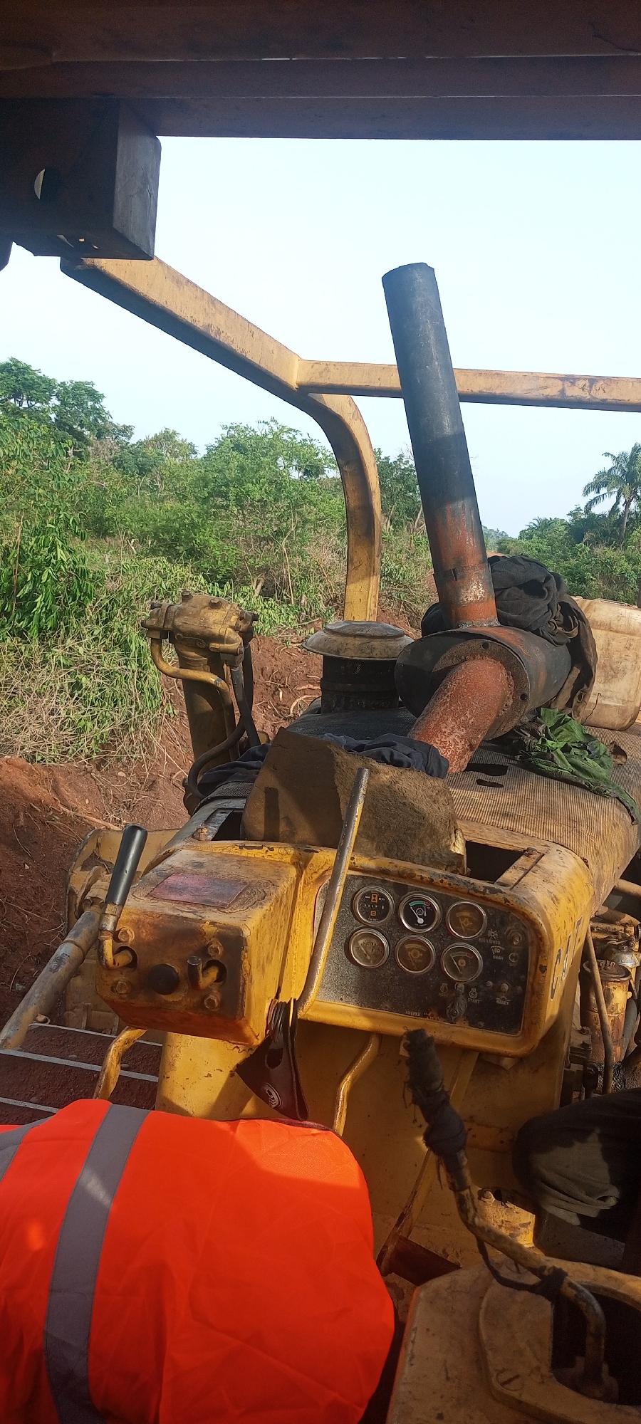 The image provides an eye-level view from within heavy yellow construction machinery, likely a bulldozer or track-type tractor, operating in an outdoor, rural setting in Otunade, Nigeria.
In the lower left foreground, a bright orange high-visibility safety vest with grey reflective stripes is draped over a component of the machine, suggesting operator presence or recent activity.
The midground is dominated by the well-used, weathered yellow machinery. A control panel is visible, featuring several circular analog gauges that appear to monitor functions like fuel, temperature, and pressure. Below these, various toggle switches are present, with visible labels including "HEAD LIGHTS," "START," "RESET," and "BRAKE." Multiple control levers with black grips are positioned around the panel, indicating operational controls for the machine. A prominent, rusty, dark-colored exhaust pipe rises vertically from the engine compartment, with dark fabric materials draped around its base. The robust yellow metal framework of the machine's roll-over protective structure (ROPS) or canopy extends above and around the operator's area.
The background showcases a vibrant, green landscape characterized by dense vegetation, including numerous trees and shrubs, consistent with a tropical environment. The ground immediately behind the machinery is reddish-brown earth, indicating recent disturbance or excavation. Further in the distance on the right, taller trees, possibly palm trees, dot the horizon. The sky is bright and clear, suggesting a daytime scene, possibly during the morning or late afternoon. The overall scene depicts heavy equipment at a worksite, likely involved in land clearing or earthmoving.