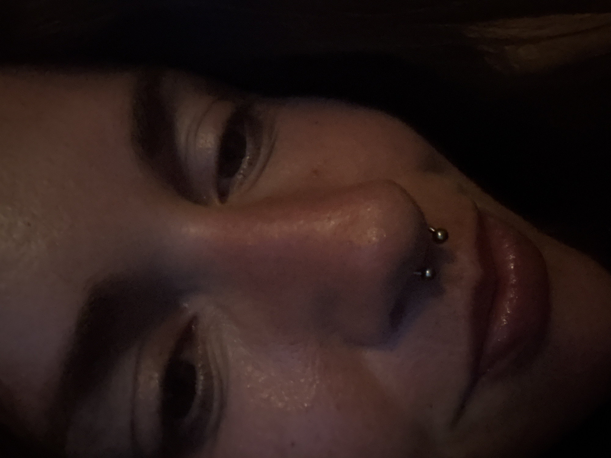 A close-up, dimly lit, underexposed photograph shows the lower face of a person, with a focus on their nose and lips. The subject has a septum piercing with two small silver balls. Their lips appear glossy, suggesting the application of lip balm or gloss. The lighting creates strong shadows, obscuring details of the eyes and the upper part of the face. The background is dark and indistinct. The image's setting cannot be determined from the visual information provided.