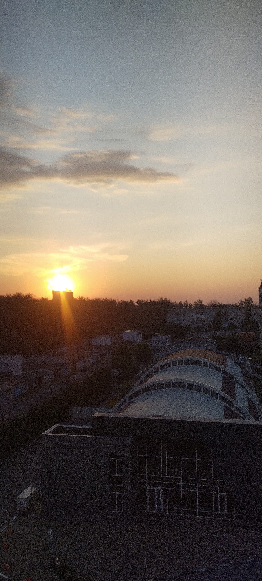 A vertical, slightly grainy photograph captures a city skyline at sunset. The sky is a gradient of pale blue at the top, transitioning to warm oranges and yellows as it nears the horizon, indicating the sun is setting. A few scattered clouds are visible in the sky.
Below the sky, a dark band of trees forms a silhouette against the sunset. Behind the trees, the tops of various buildings are visible, including what appears to be apartment buildings and smaller structures. The sun itself is a bright, intense source of light peeking from behind a taller building, casting rays that fan out downwards.
In the foreground, a modern, dark-colored building with a large, angular glass facade dominates the lower part of the frame. To the left of this building, a smaller, light-colored rectangular structure, possibly a utility box or storage unit, sits on the ground. The ground appears to be asphalt, with faint white lines suggesting a parking lot or road. Orange traffic cones are visible near the foreground building. Further back and to the right, a large, curved roof structure with visible supporting ribs is partially visible, also appearing to be a building. The overall scene suggests an urban environment at dusk.