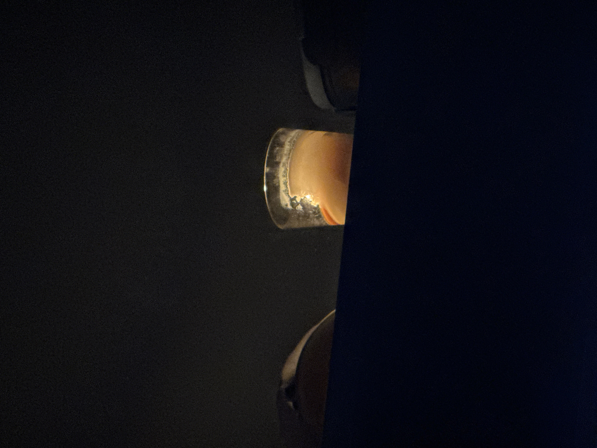 The image shows a close-up, low-lit scene. Partially visible are two items. A glass, possibly a drinking glass, containing a light-colored liquid and some foam around the rim is present. Below that is a human head. The background is dark and out of focus. No information suggests this image was taken in Izhevsk, Russia.