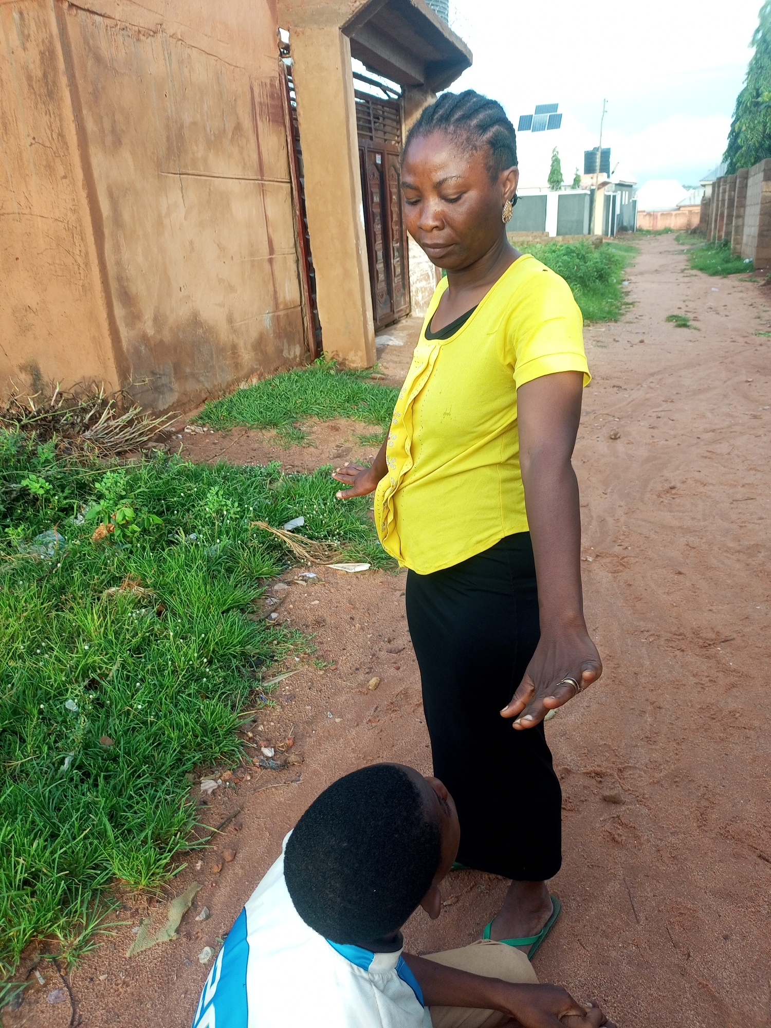 In Tempure, Nigeria, a young boy kneels in the dirt, looking up at a woman standing over him. The woman, wearing a bright yellow t-shirt and a black skirt, gestures with her hands. She has her hair braided and wears gold earrings. The boy is dressed in a white and blue shirt and tan pants. They are in an outdoor environment with a dirt path, dry grass, and a textured, ochre-colored wall behind them. In the background, several buildings and trees are visible. The sky is overcast.